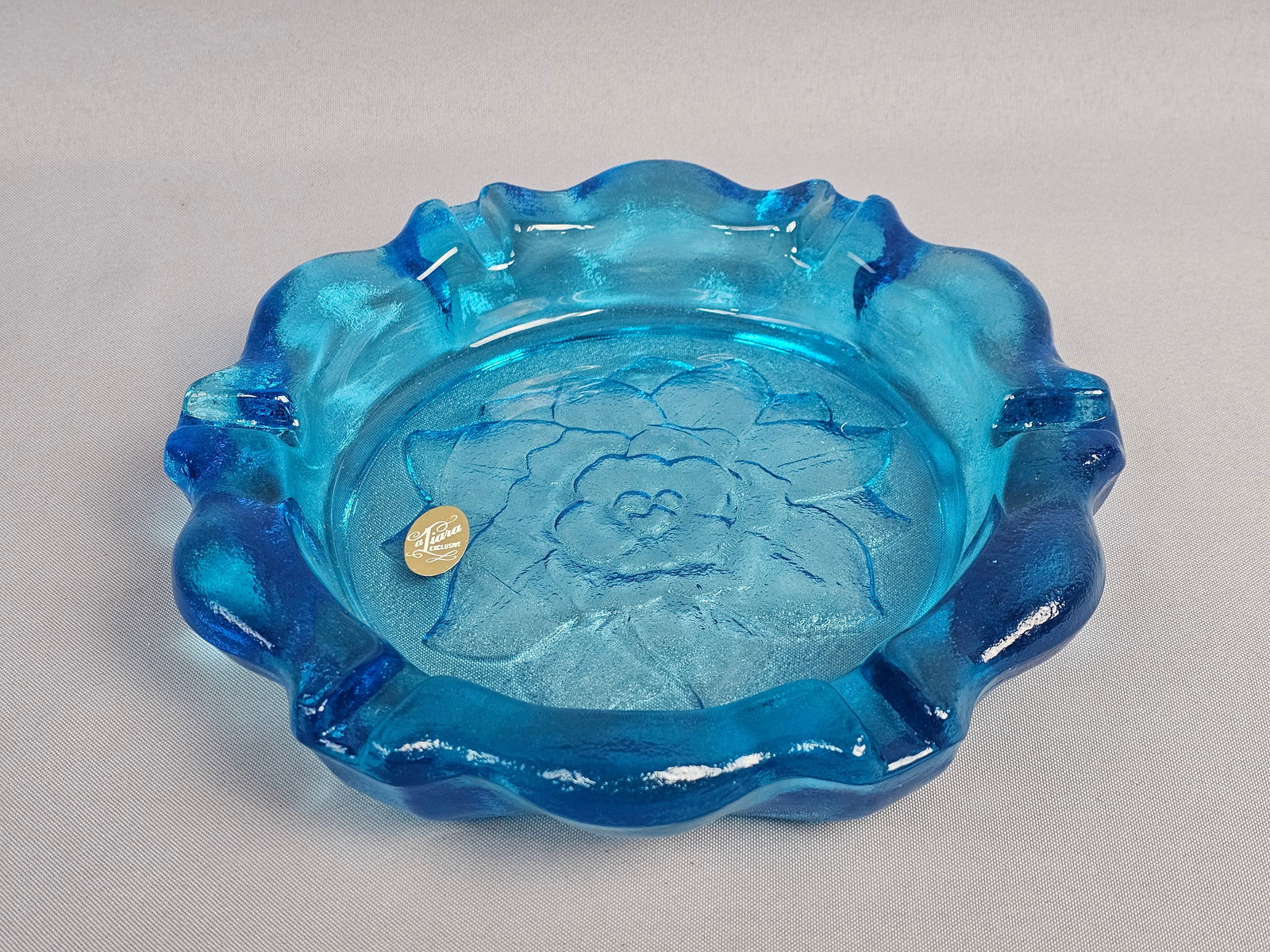 Tiara Exclusives Pressed Glass Ashtray: Produced for Tiara Exclusives by Indiana Glass, this vintage ashtray is crafted from pressed glass in a vibrant translucent color known as Bermuda Blue. The design features a wide scalloped rim with f