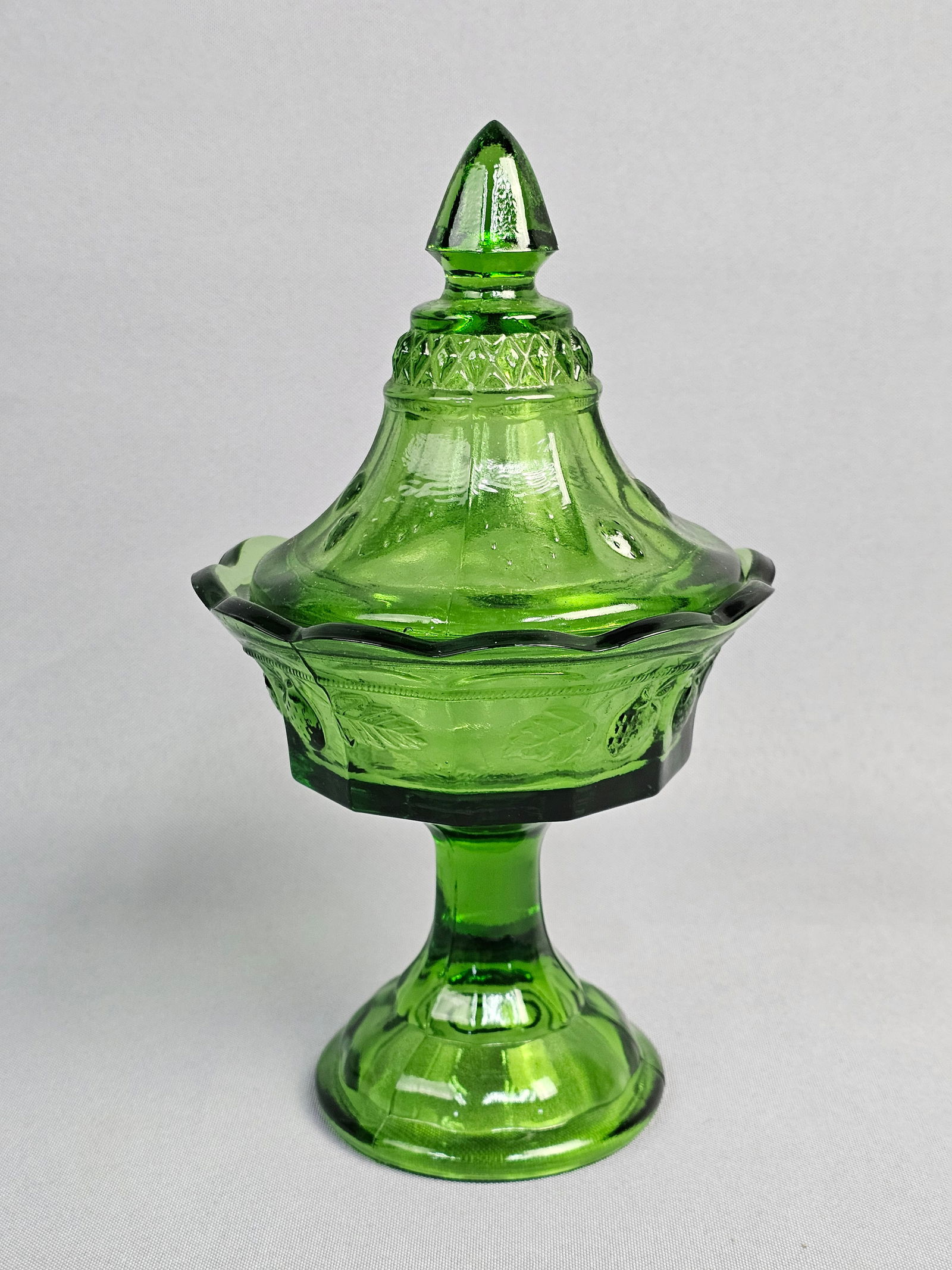 Indiana Glass Pressed Glass Emerald Green Covered Compote: This two-piece covered compote is crafted from vibrant, transparent emerald green pressed glass. The design is attributed to Indiana Glass, featuring their Strawberry pattern with embossed fruit and l