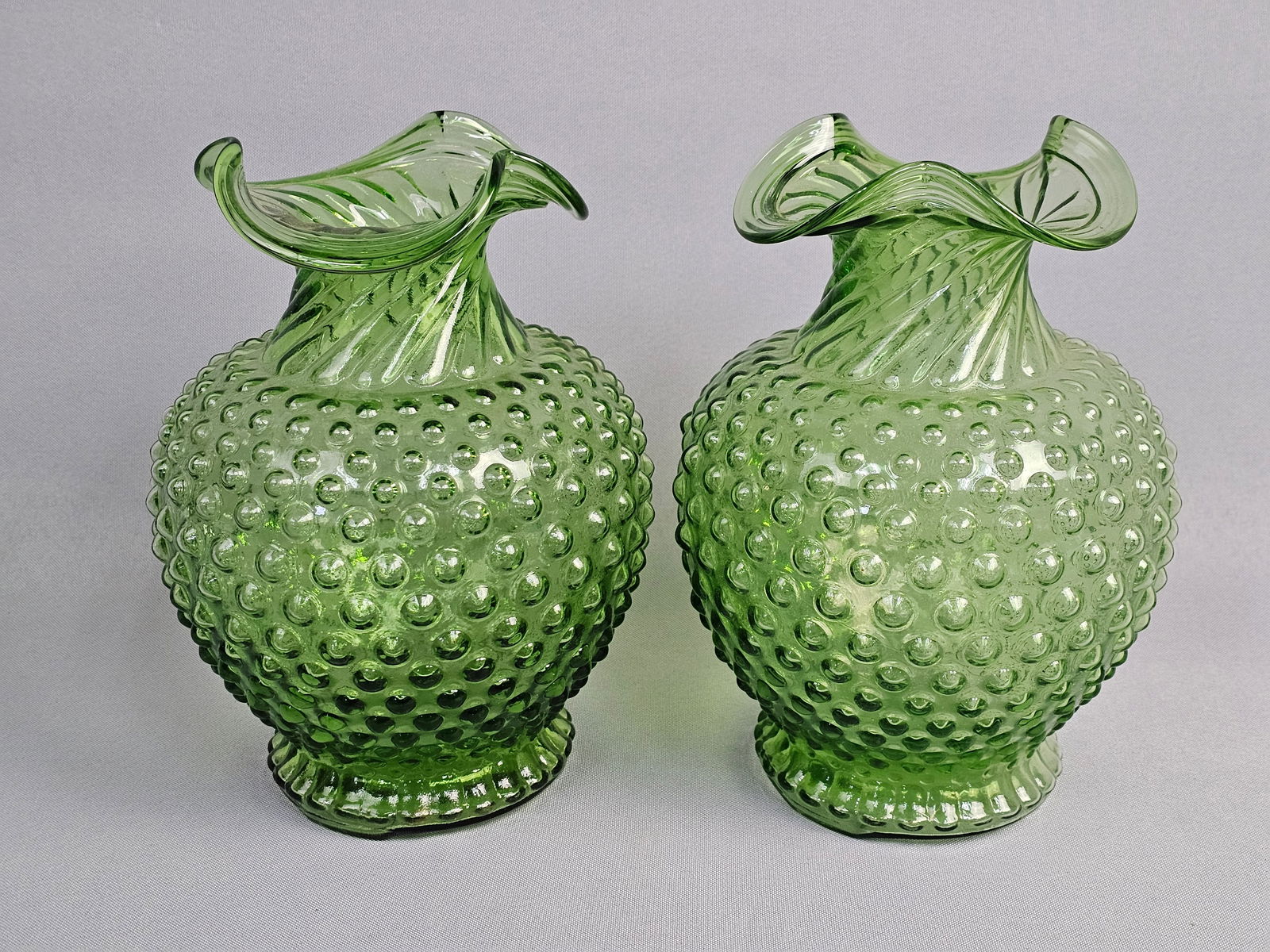 A Pair of Art Glass Hobnail Vases: This pair of mid-century art glass vases is rendered in a vibrant transparent emerald green. The vases were created using a combination of mold-blown and hand-finishing techniques; the neck displays a