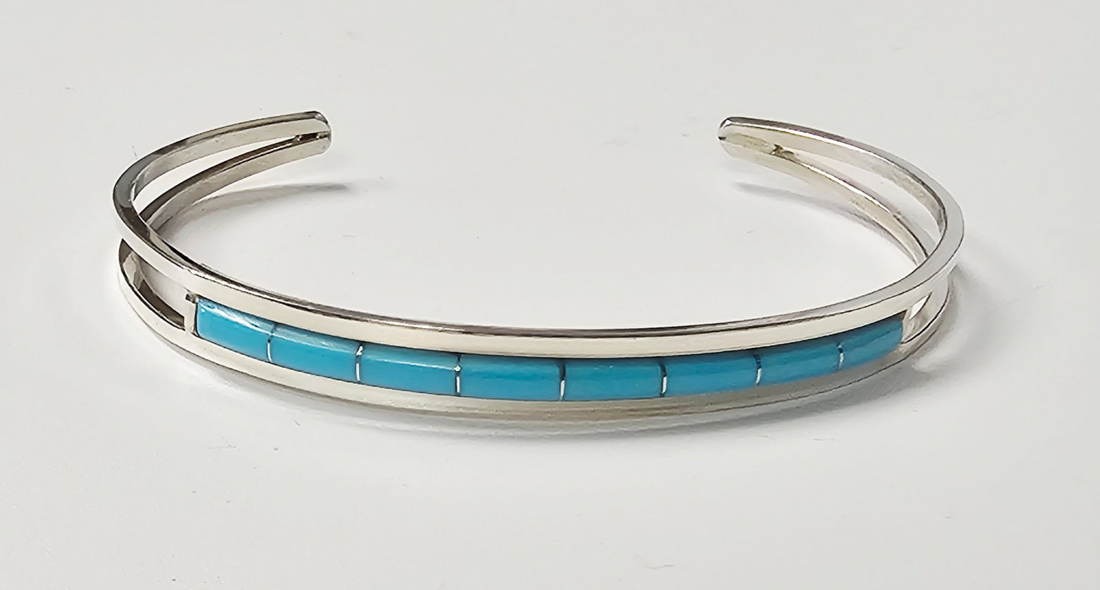 Running Bear Sterling Turquoise Cuff Bracelet 8.7 Grams: This sterling silver cuff bracelet features a split-shank design with a central channel-set inlay. The inlay consists of eleven rectangular, bright blue stones characteristic of turquoise. The piece i