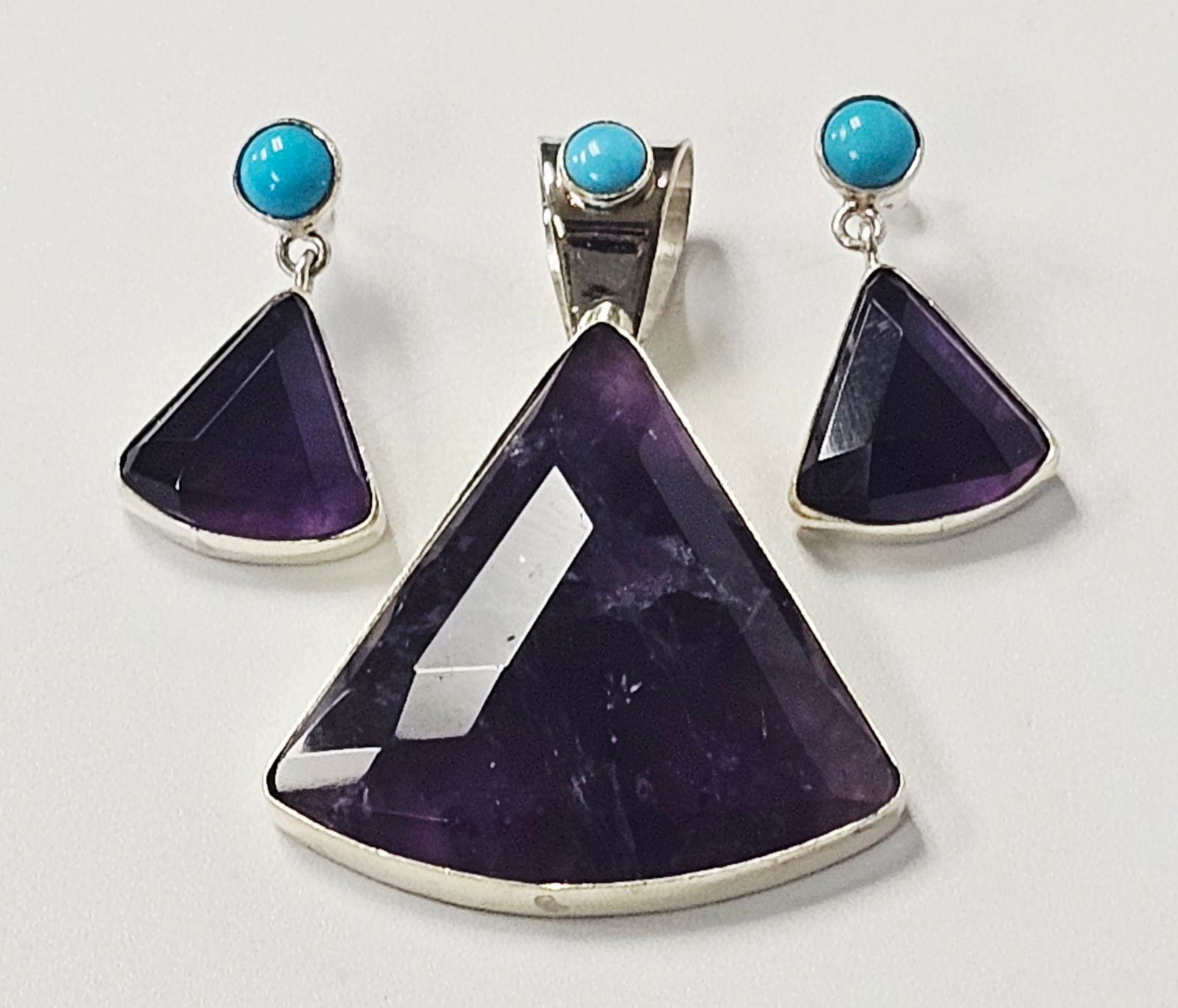 Sterling Amethyst & Turquoise Earrings & Pendant 27.0 Grams: This jewelry set consists of one pendant and one matched pair of drop earrings. The pieces are crafted from sterling silver and feature faceted trilliant-cut purple stones, likely amethyst, and round