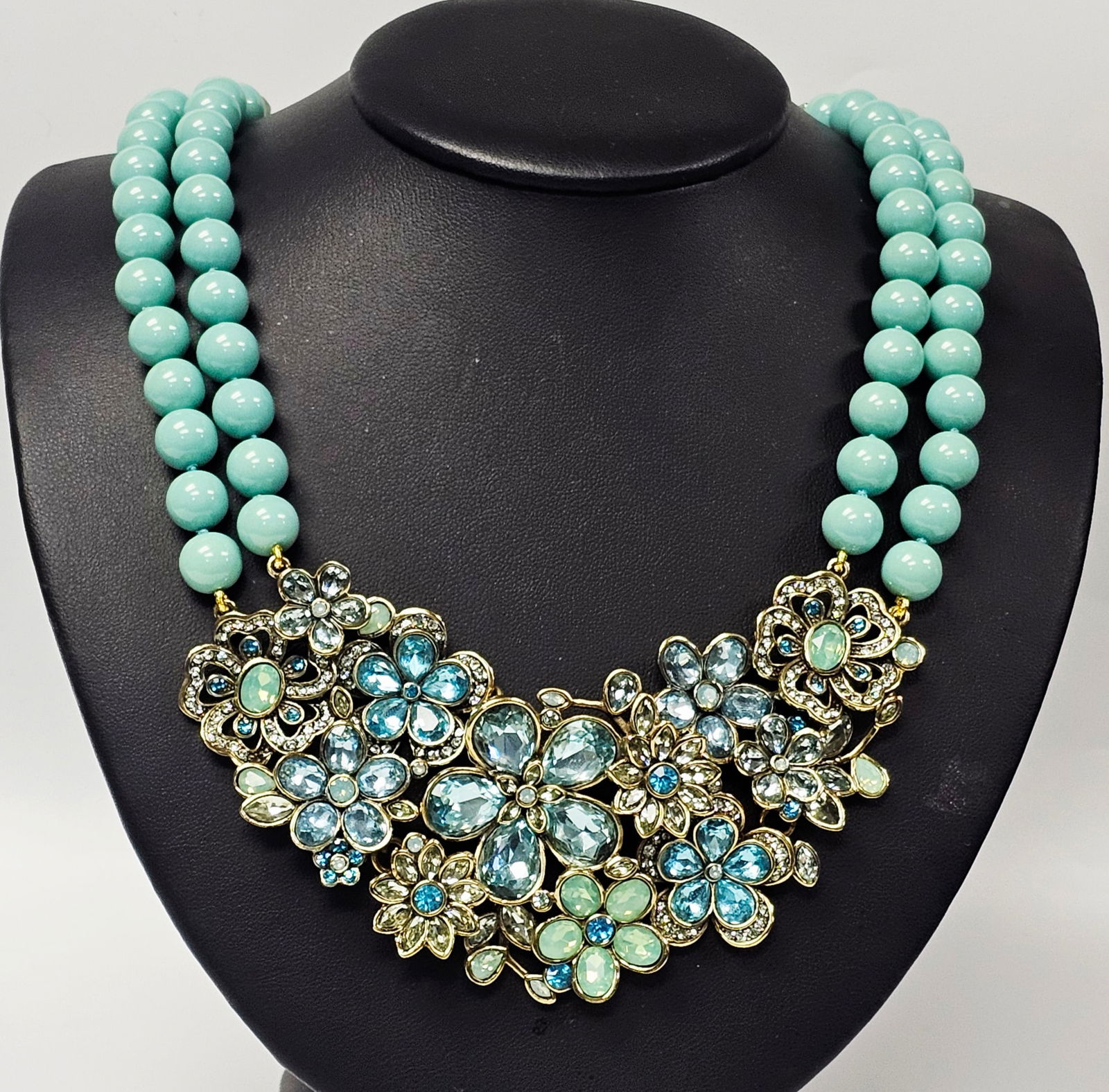 Heidi Daus Floral Crystal Bib Necklace with Turquoise Beads: A Heidi Daus bib necklace featuring two strands of turquoise-colored beads and a gold-toned floral centerpiece set with blue and green crystals, measuring approximately 5 inches across the bib. Measur
