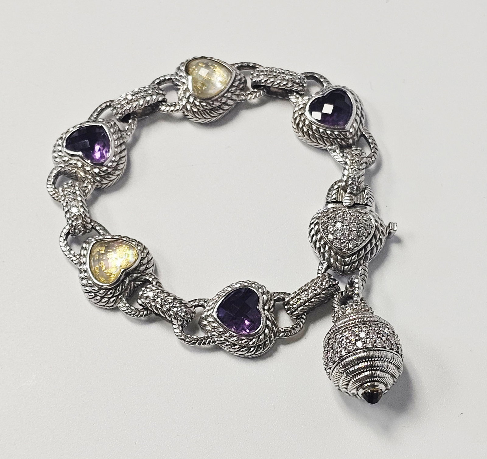 Judith Ripka Sterling Cubic Zirconia Bracelet 54.7 Grams: A sterling silver heart-link bracelet by Judith Ripka featuring five heart-shaped stations. Three stations are set with faceted purple stones, and two are set with yellow stones containing gold-colore