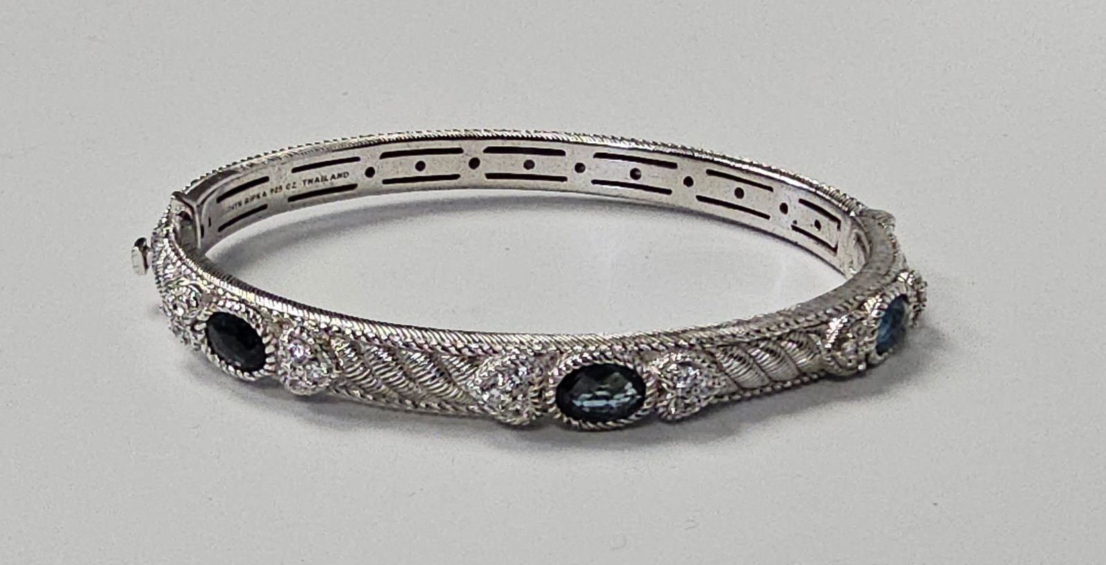 Judith Ripka Sterling Hinged Bangle Bracelet 31.7 Grams: A sterling silver hinged bangle bracelet by Judith Ripka. The piece features an ornate cable-textured band set with three oval-faceted dark blue gemstones. These are interspersed with clusters of smal