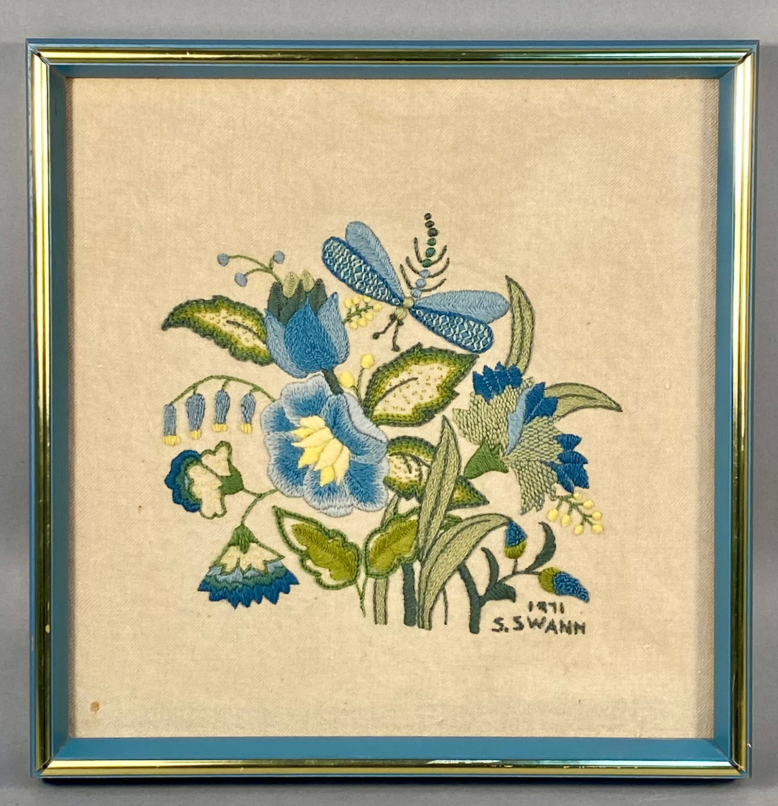 Framed Erica Wilson Floral Crewel Embroidery Signed S. Swann 1971: A framed crewel embroidery piece featuring a stylized floral arrangement with a central blue dragonfly. The work is executed in wool thread on a light-colored linen or cotton-blend ground fabric. The