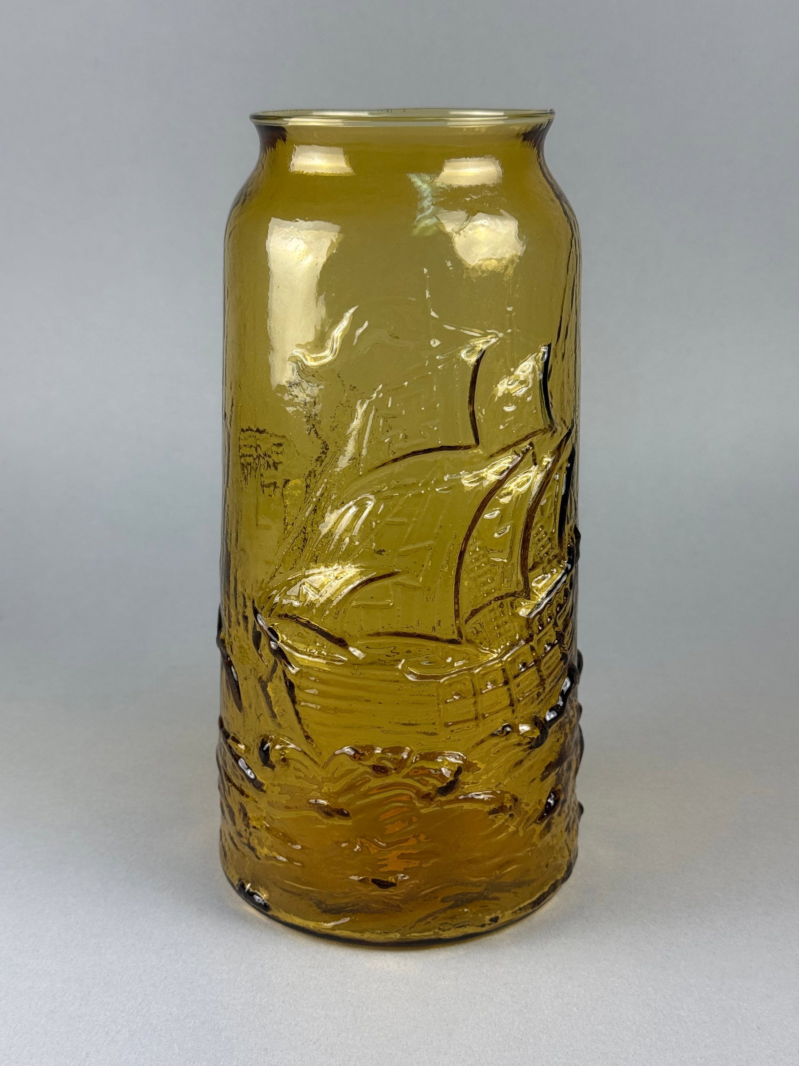 An Amber Pressed Indiana Glass Vase with Relief-Molded Ship and Bird Decoration: A cylindrical amber-toned pressed glass vase featuring relief-molded decorative motifs. The body is ornamented with a sailing ship on one side and a bird in flight, resembling a swan, on the reverse.
