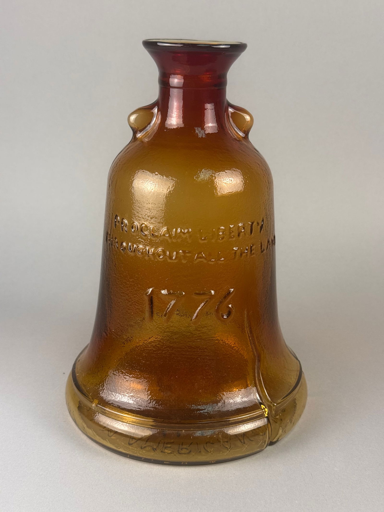 Hand Blown Amber Glass Liberty Bell Decanter, 1976: A K.L.M. American Classic hand-blown glass decanter in the form of the Liberty Bell. The vessel features a textured amber glass body that transitions to a ruby red tone at the flared neck. The body is