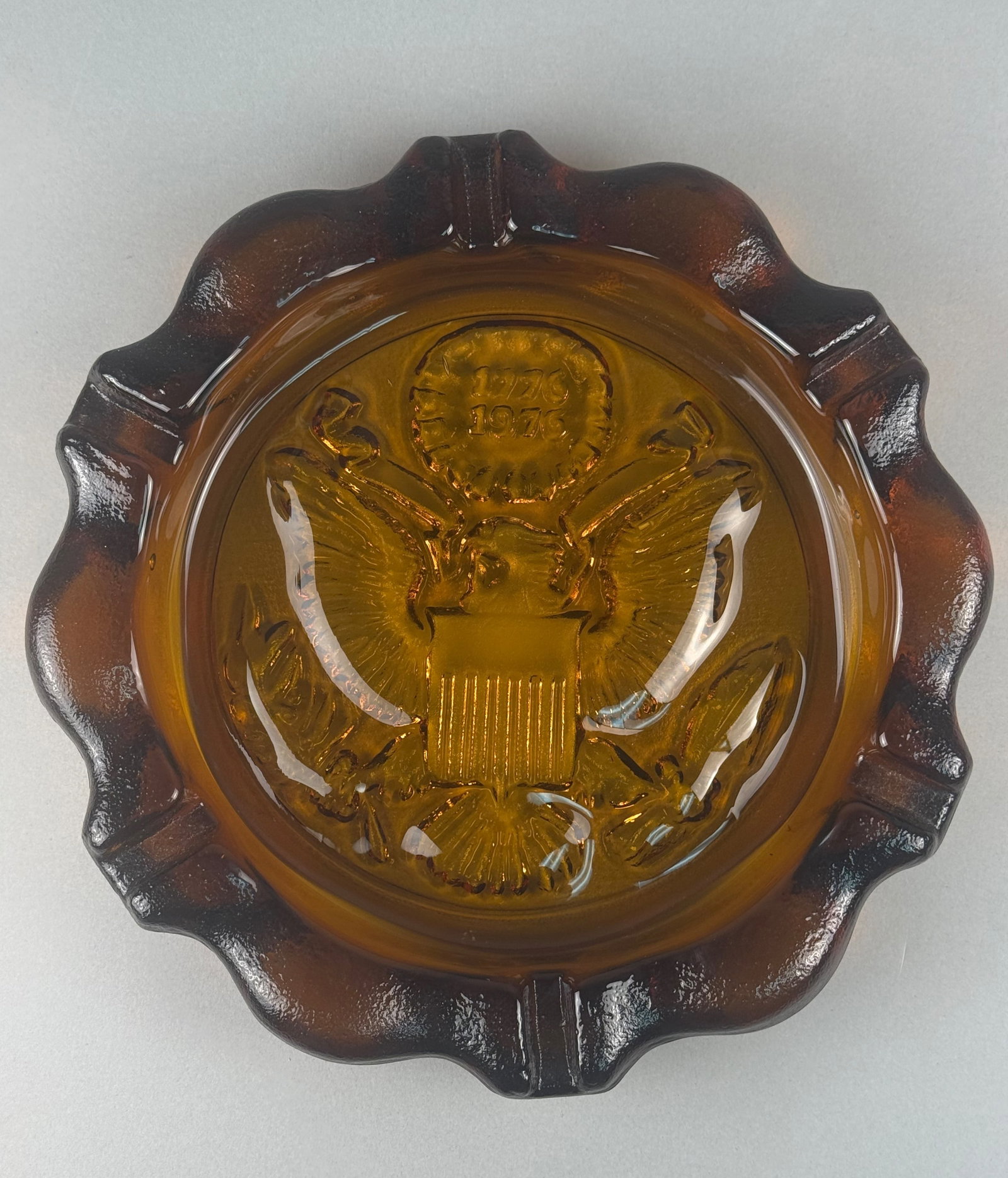 Vintage Amber Pressed Glass Bicentennial Commemorative Eagle Ashtray: A vintage commemorative ashtray produced for the United States Bicentennial, attributed to Indiana Glass for Tiara Exclusives. This circular vessel is crafted from heavy pressed glass in a warm honey-
