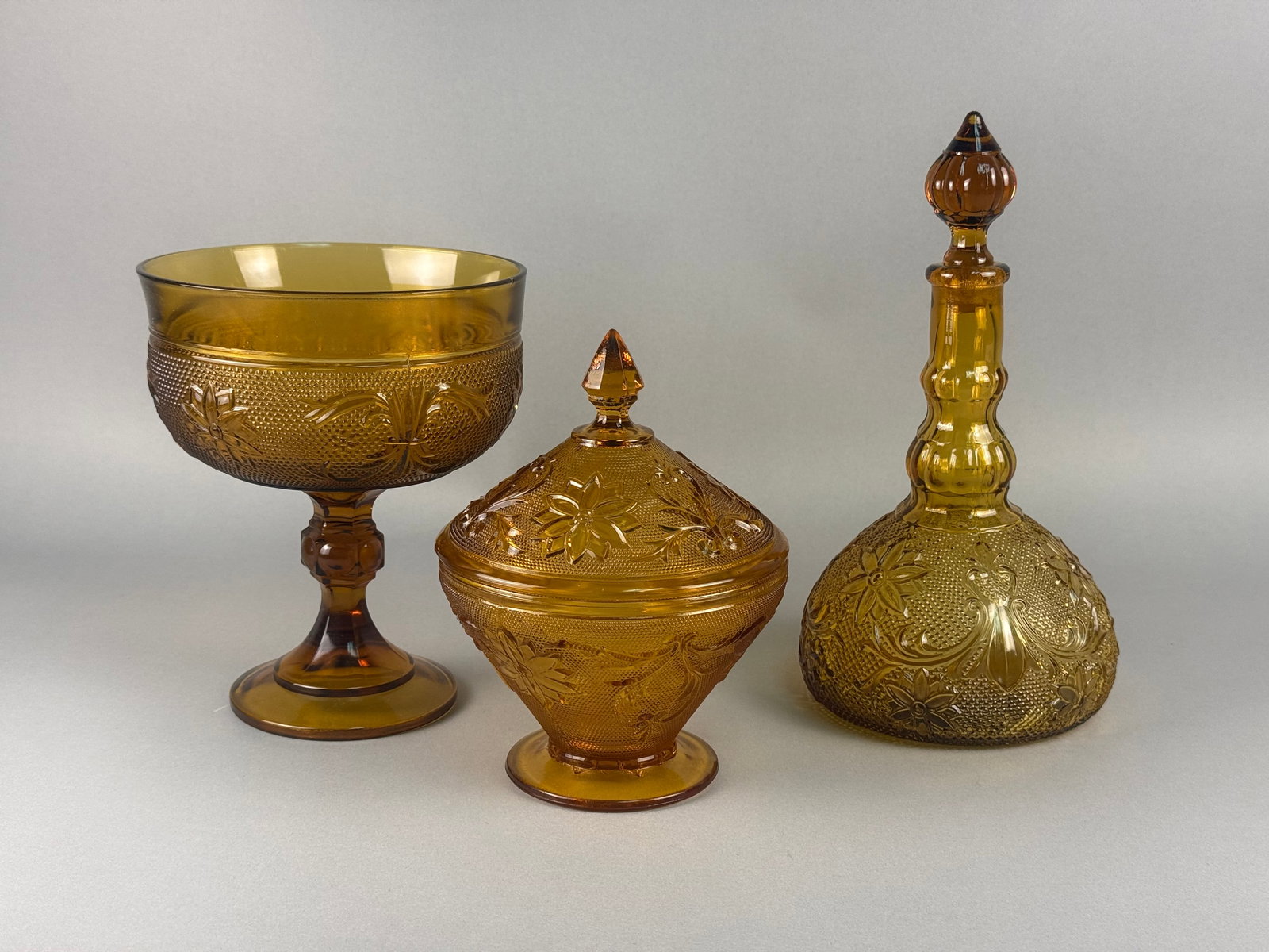 A Trio of Amber Pressed Glass Tableware in the Sandwich Pattern: A group of three amber-colored pressed glass items in the Sandwich pattern, attributed to Tiara Indiana Glass. The collection consists of a pedestal bowl with a flared rim, a covered candy dish with a