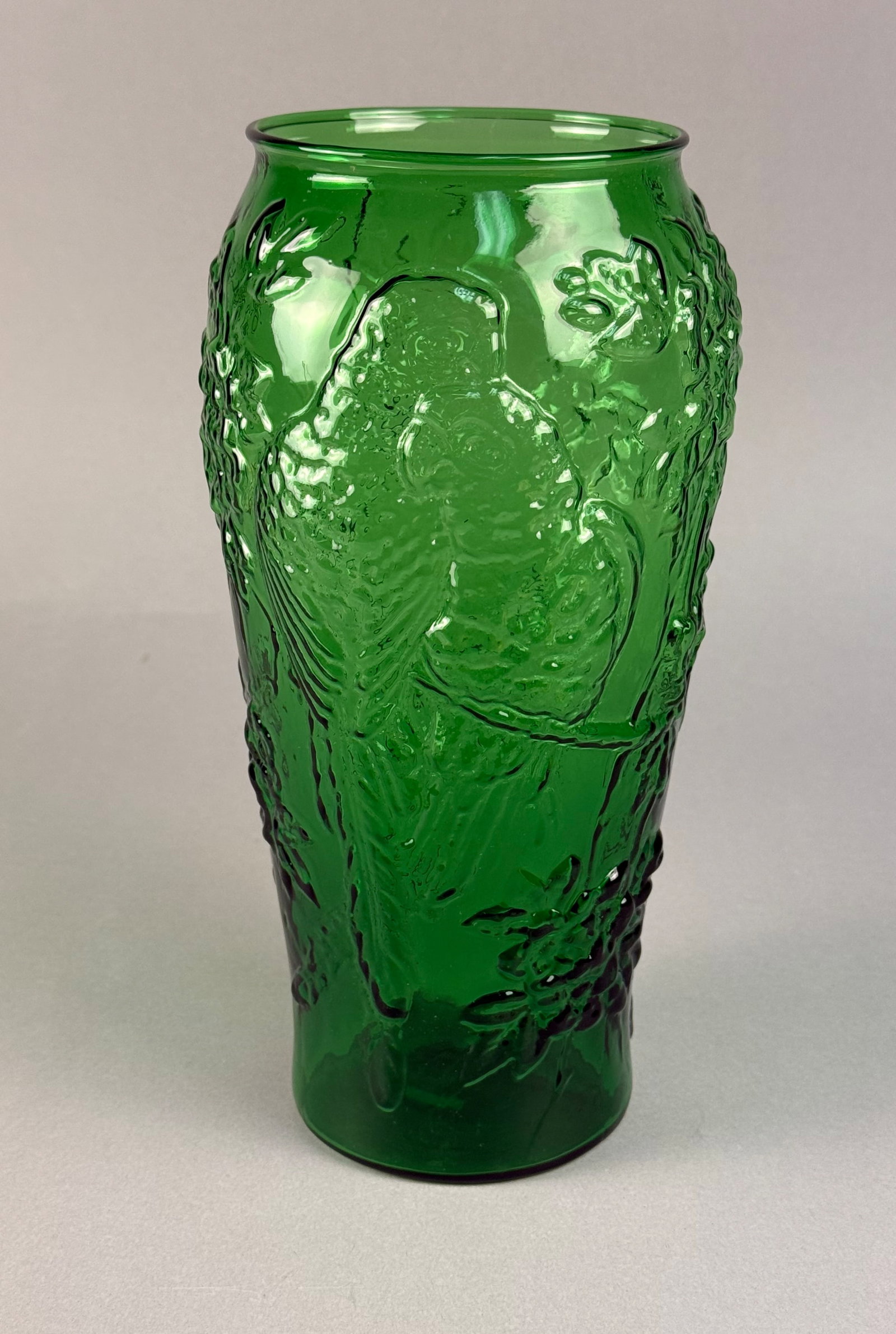 A Tiara Exclusives Indiana Glass Emerald Green Textured Parrot Vase: A pressed glass vessel in emerald green, attributed to the Indiana Glass Company. The vase features a tapered cylindrical body with a relief-molded design depicting two parrots surrounded by dense flo