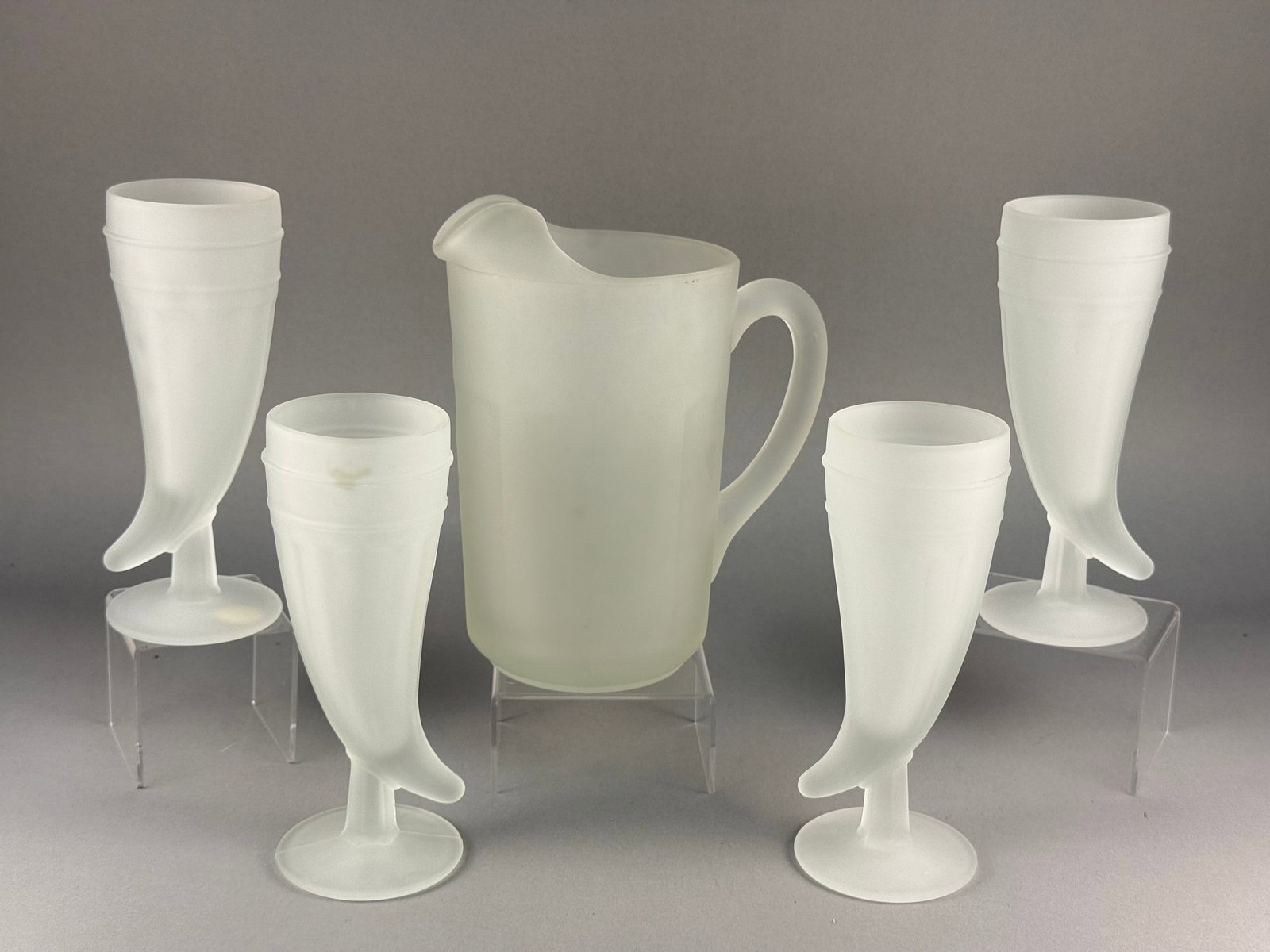 A Tiara Exclusive Frosted Glass Pitcher and Powderhorn Tumbler Set: A set of white frosted glass beverage ware by Tiara Exclusive. The collection includes a cylindrical pitcher and several tumblers fashioned in the form of powder horns. The pitcher is designed with a