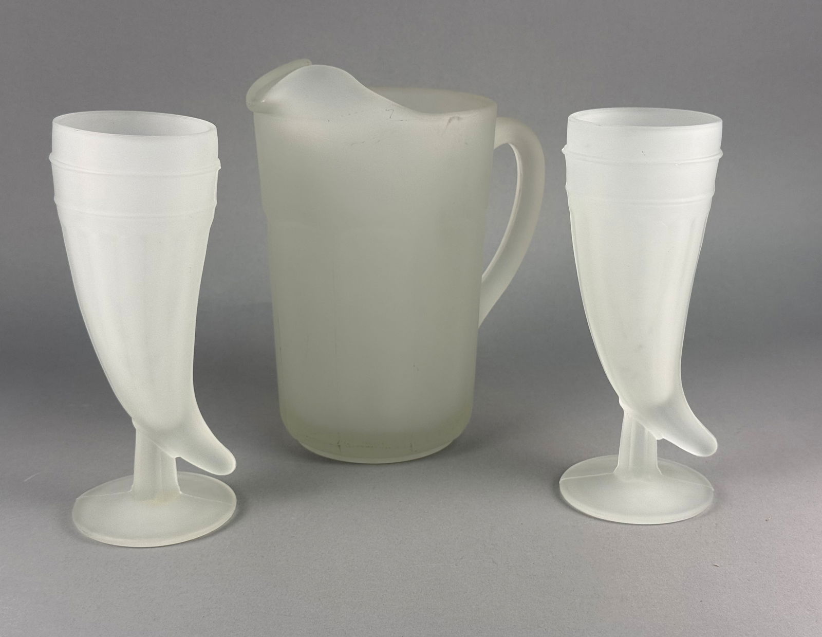 Vintage Frosted White Plastic Pitcher and Horn Goblet Set: A three-piece beverage set comprising one pitcher and two footed goblets, crafted from molded translucent white plastic with a frosted or satin-style finish. The pitcher features a faceted body with a