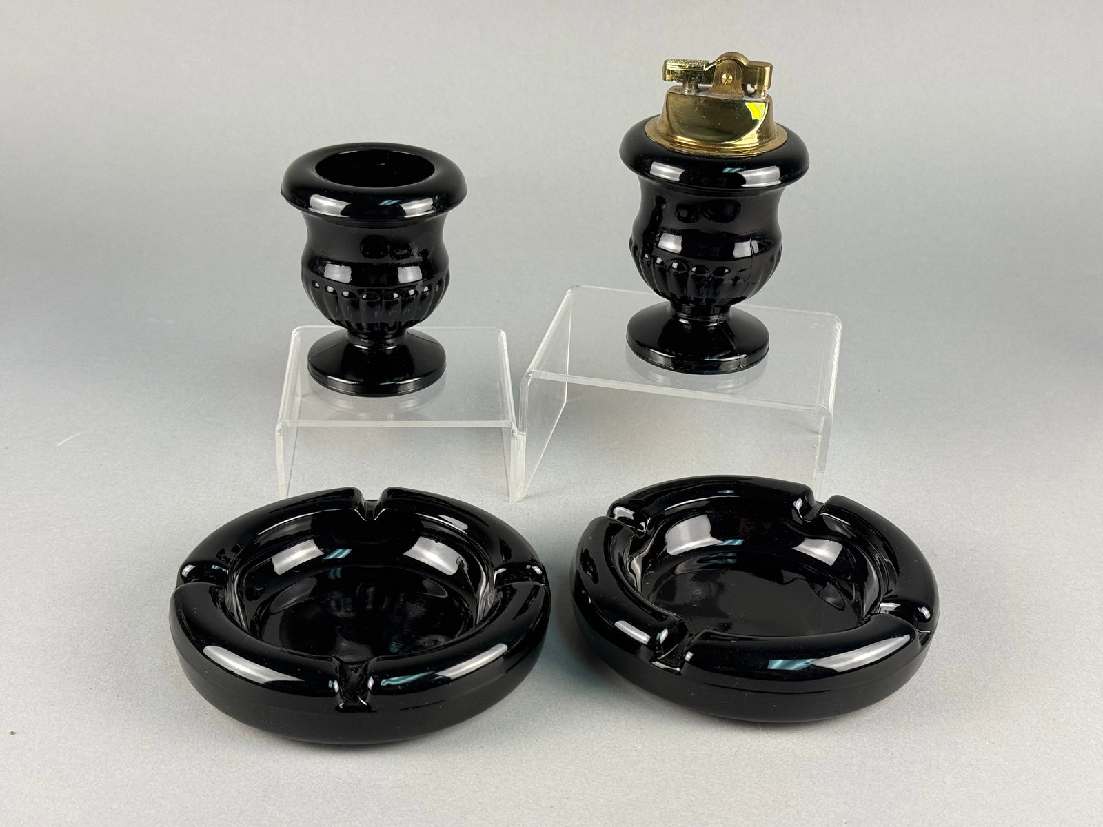 A Set of Black Opaque Glass Smoking Accessories From Tiara Exclusives: This set of black opaque glass smoking accessories includes circular ashtrays and pedestal urn-shaped vessels. The glass is a deep opaque black with a high-gloss finish. The urn-shaped vessels feature