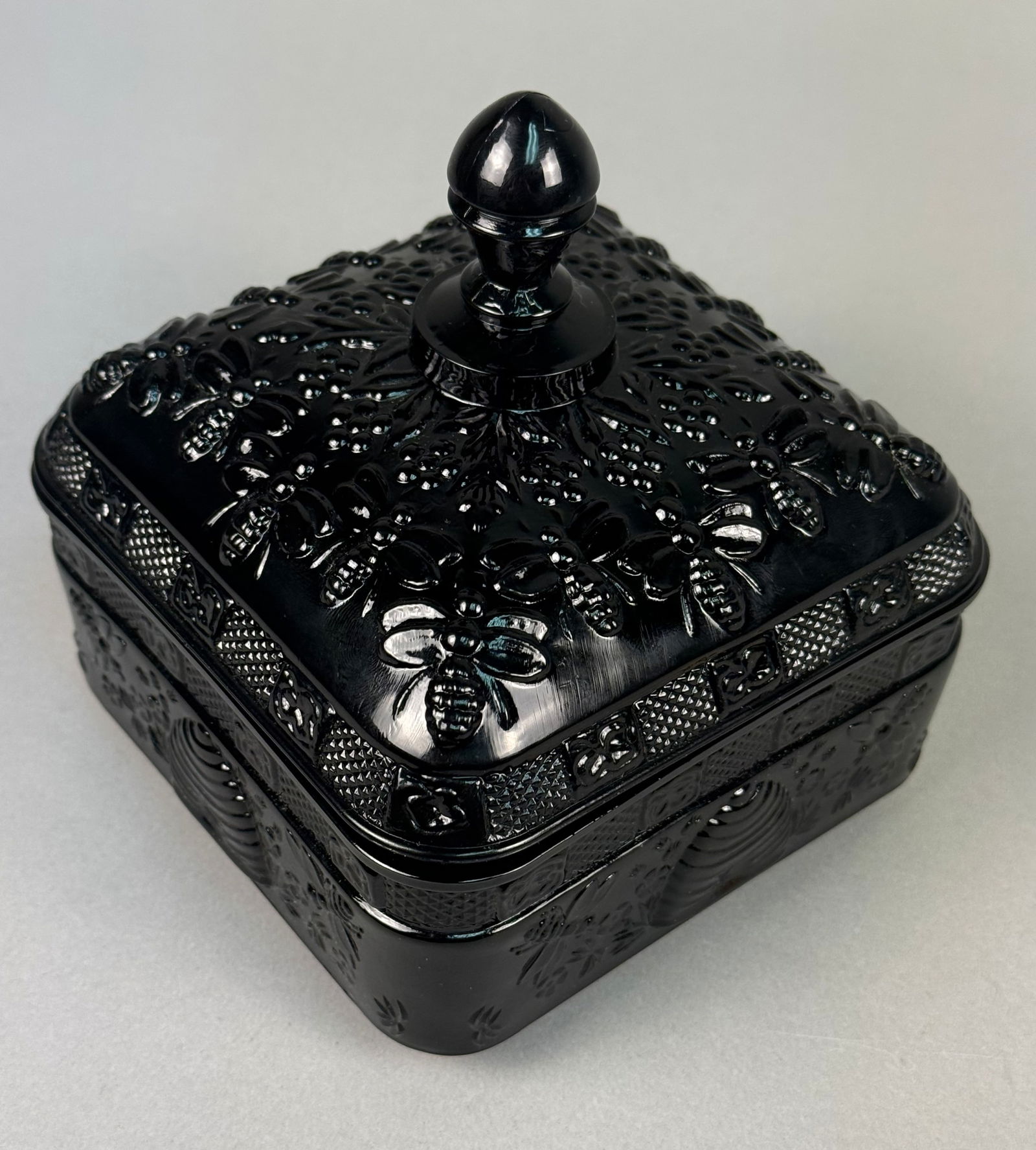 Tiara Exclusives Black Glass Honey Bee Pattern Square Covered Dish: This square covered dish is crafted from opaque black pressed glass in the Honey Bee pattern, attributed to Tiara Exclusives and manufactured by Indiana Glass. The lid features a central bulbous finia