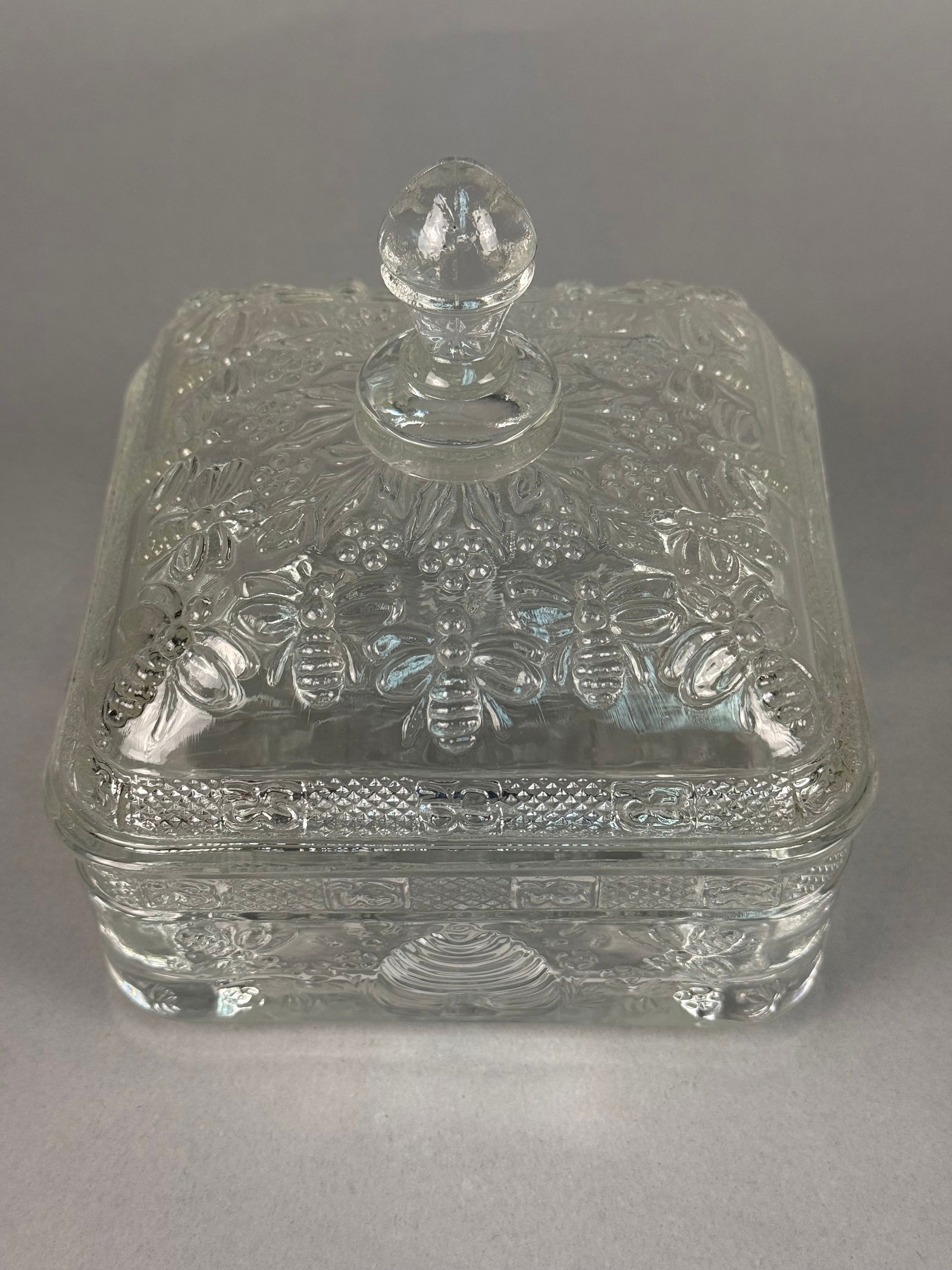 Clear Pressed Glass Footed Lidded Box in the Beehive and Honeybee Pattern: This square, footed clear pressed glass lidded box is in the Beehive and Honeybee pattern, attributed to Indiana Glass for Tiara. The piece is constructed of heavy pressed glass with high-relief molde