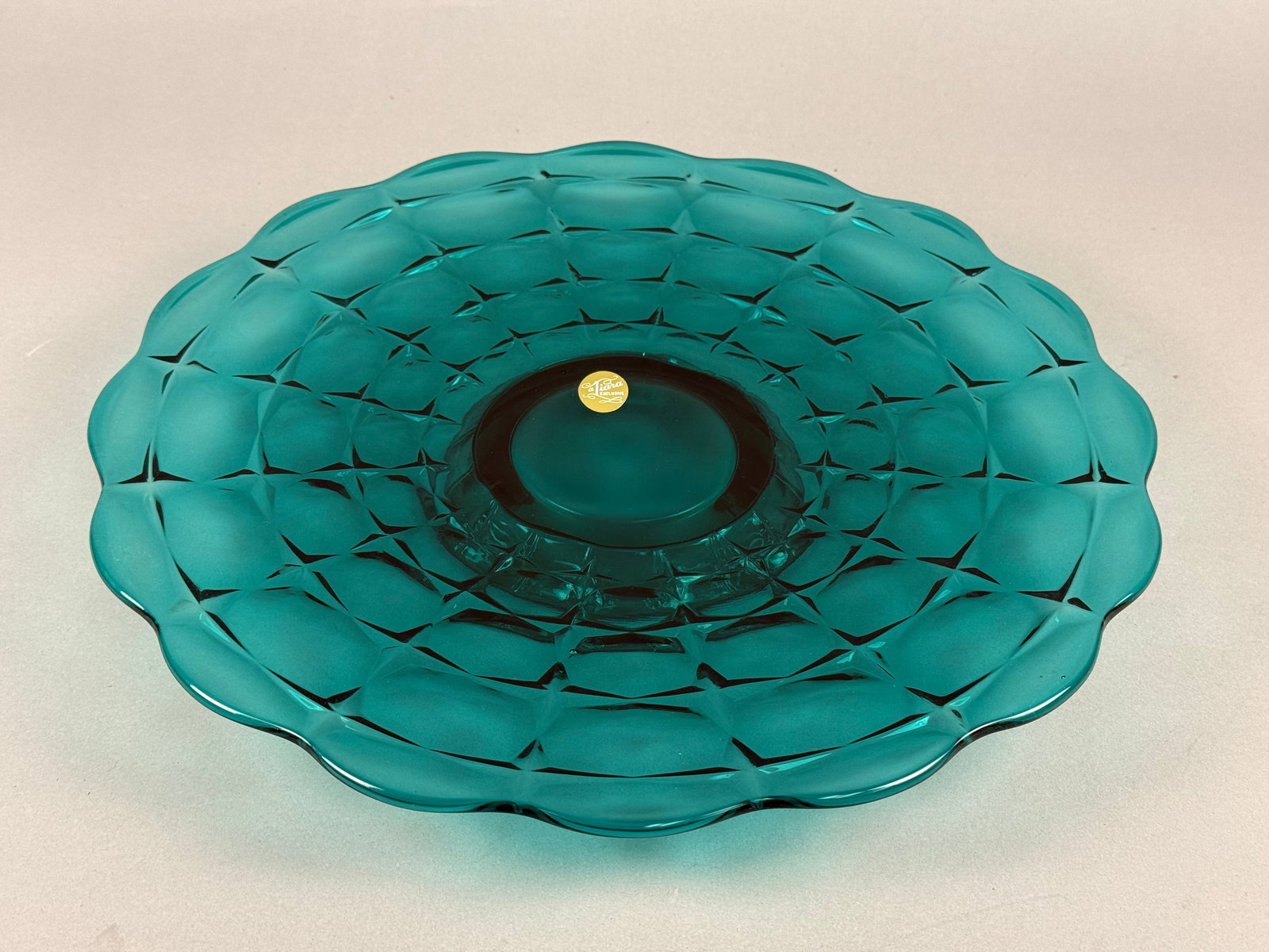 Tiara Exclusives Constellation Pattern Teal Blue Glass Cake Stand: A Tiara Exclusives pressed glass cake stand in the Constellation pattern. The piece is made of teal blue glass and features a broad, shallow plate with a scalloped rim. The underside of the plate is m