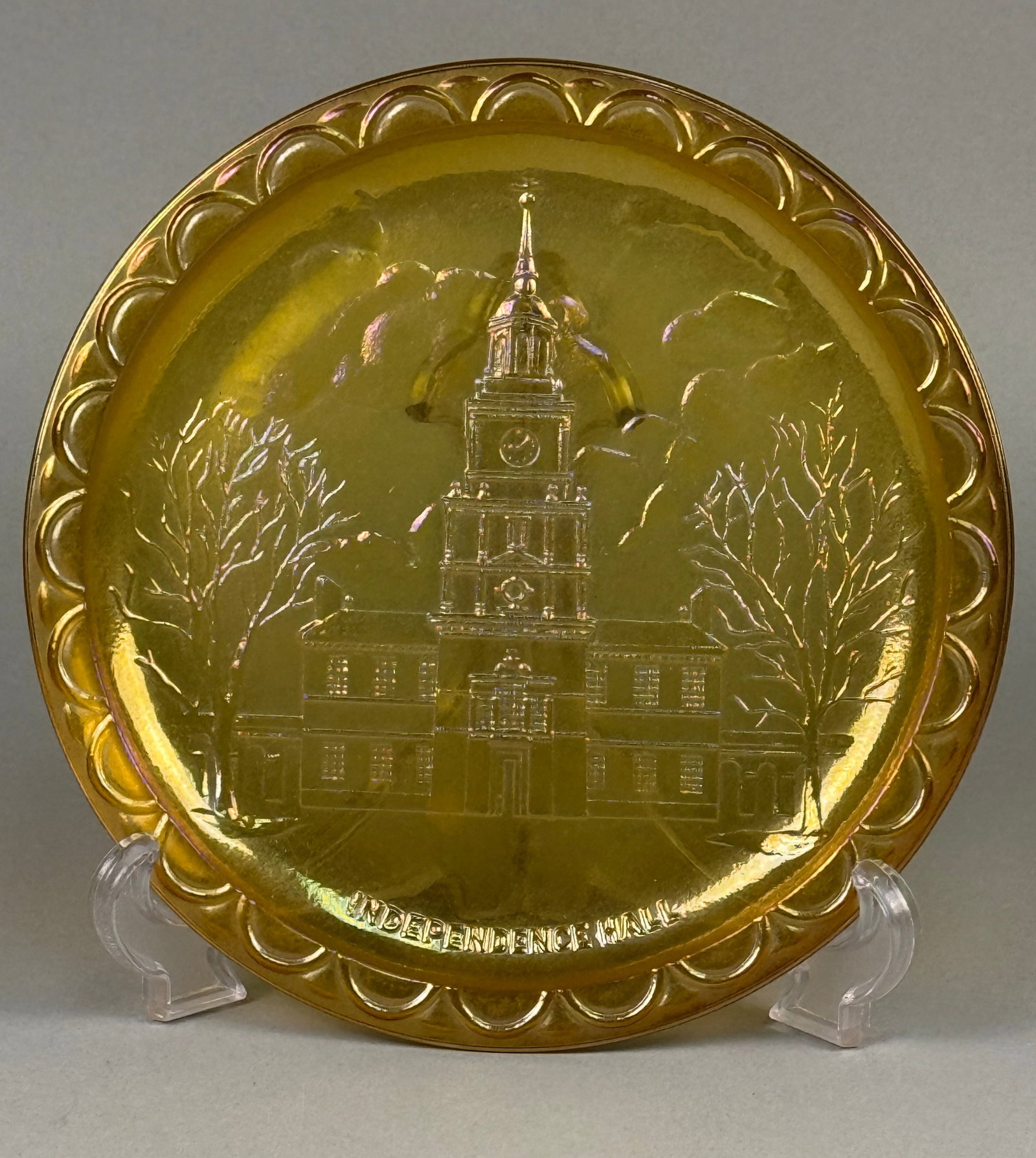 Independence Hall Commemorative Marigold Carnival Glass Plate: The item is constructed of pressed glass with a marigold iridescent surface treatment over an amber-yellow base. The central well features a raised-relief depiction of Independence Hall flanked by lea