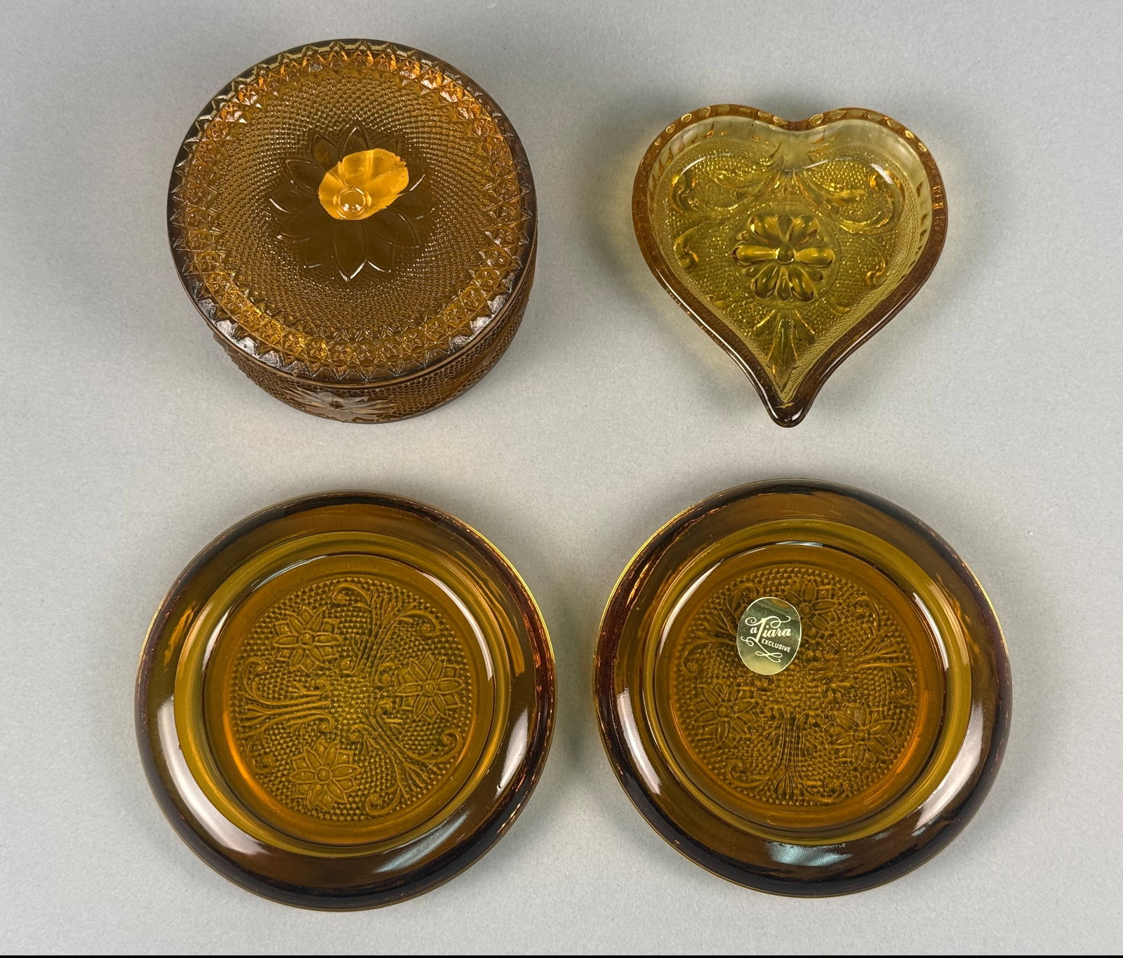 Four Pieces of Tiara Exclusives Amber Sandwich Pattern Glassware: This lot of amber pressed glassware is produced in the Sandwich pattern, featuring floral and scroll motifs with a stippled ground. The pieces are attributed to Tiara Exclusives and were manufactured