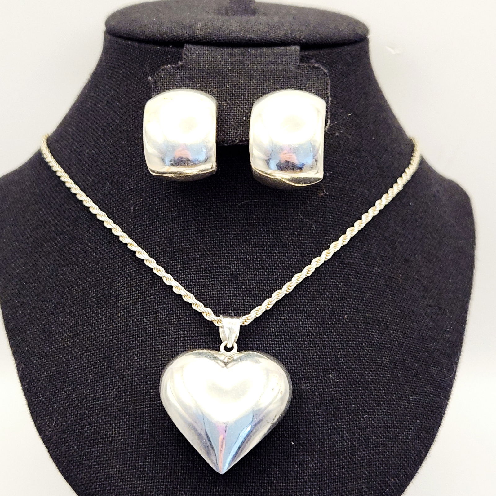 A Group of Sterling Silver Jewelry: A group of contemporary sterling silver jewelry comprising a polished heart-shaped pendant, a rope chain, and a pair of wide semi-hoop stud earrings. The pendant is a puffed heart design with a bail m