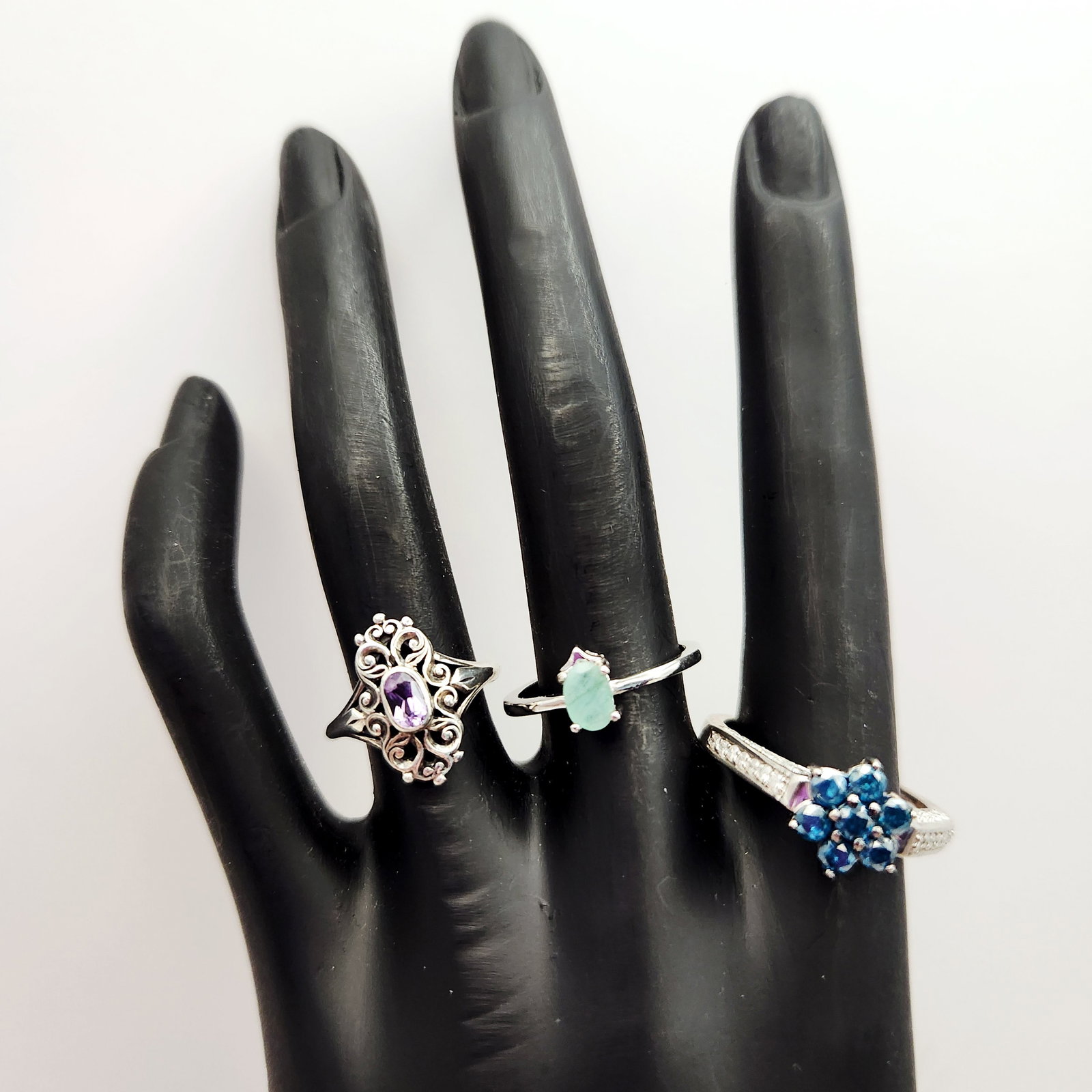 Three Gemstone Sterling Rings 8.2 Grams: This collection of rings features various gemstone-style settings. The group includes an ornate scrolling openwork mounting centering an oval-cut purple stone with illegible marks on the underside, a