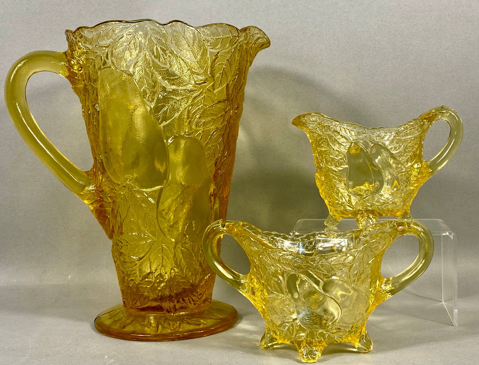 Three-Piece Yellow Pressed Glass Tableware Set in Pear Pattern: A three-piece collection of transparent yellow pressed glass tableware, comprising a tall pitcher, a creamer, and a double-handled open sugar bowl. Each vessel is decorated with a high-relief molded p