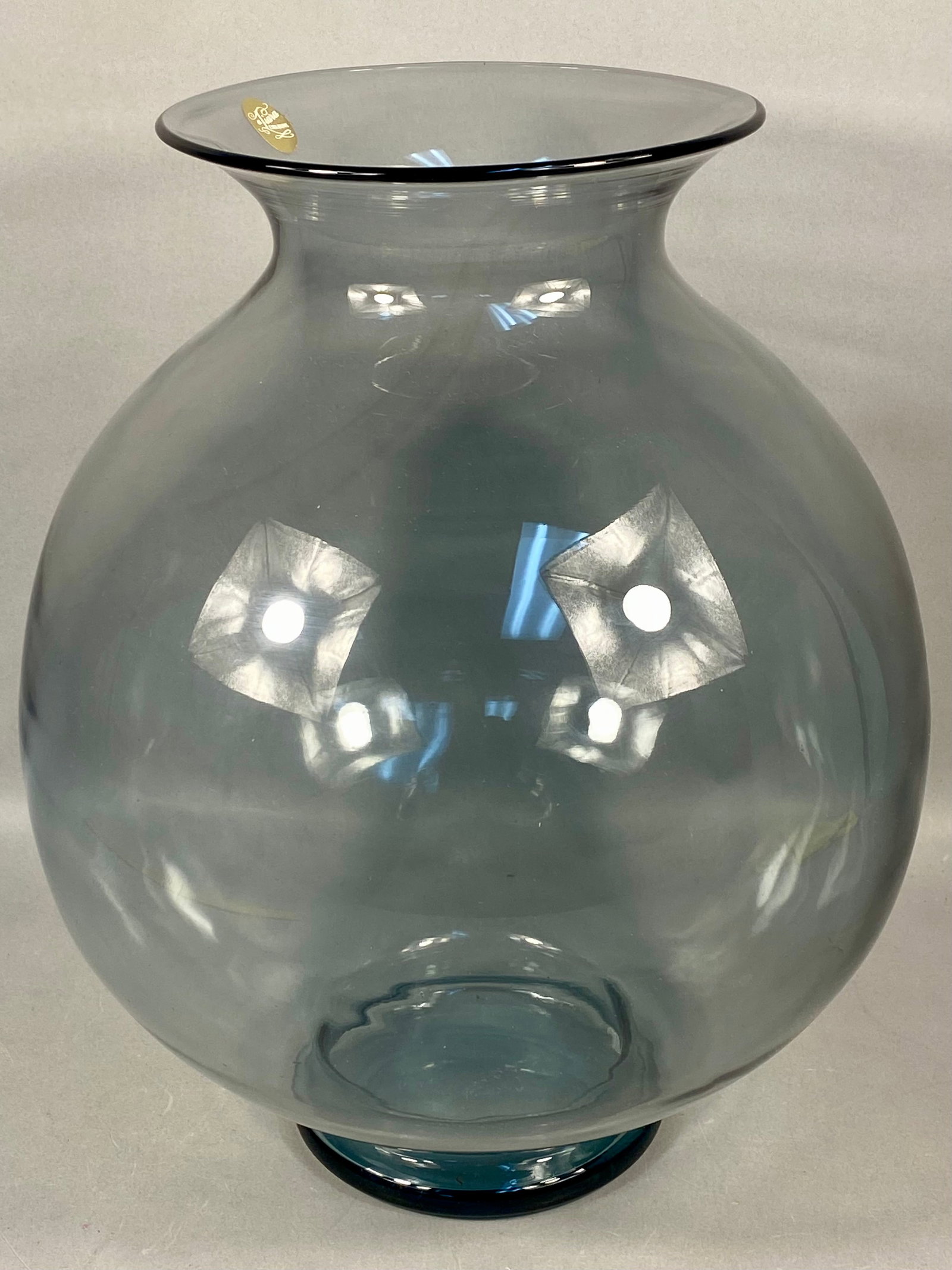 Tiara Exclusives Smoke Blue Hand Blown Glass Vase with Original Box: A large hand-blown glass vase produced for Tiara Exclusives. The vase is crafted from transparent Smoke Blue glass, characterized by a muted grayish-blue tone. The form features a rounded spherical bo