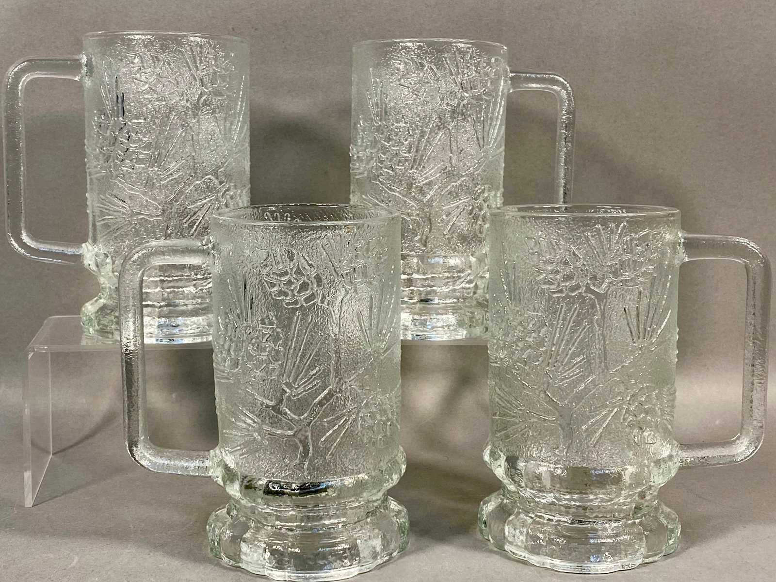A Collection of Four Tiara Ponderosa Pine Mugs: A collection of four clear glass mugs from Tiara in the Ponderosa Pine motif. Each mug features pine boughs and cones. Measurement: 5.5 inches tall Very good condition.