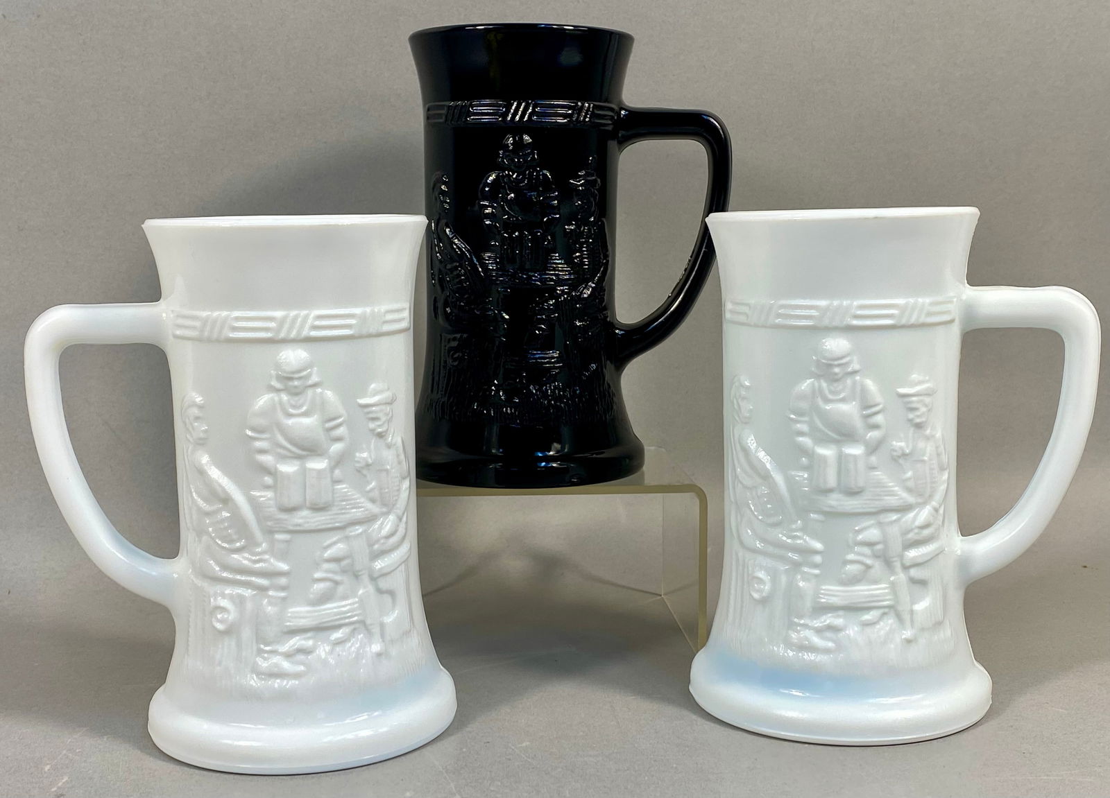 A Trio of Vintage Pressed Glass Tavern Scene Steins: This lot consists of three pressed glass steins featuring a bas-relief tavern scene. The collection includes two steins made of white opalescent milk glass and one made of opaque black glass. Each pie