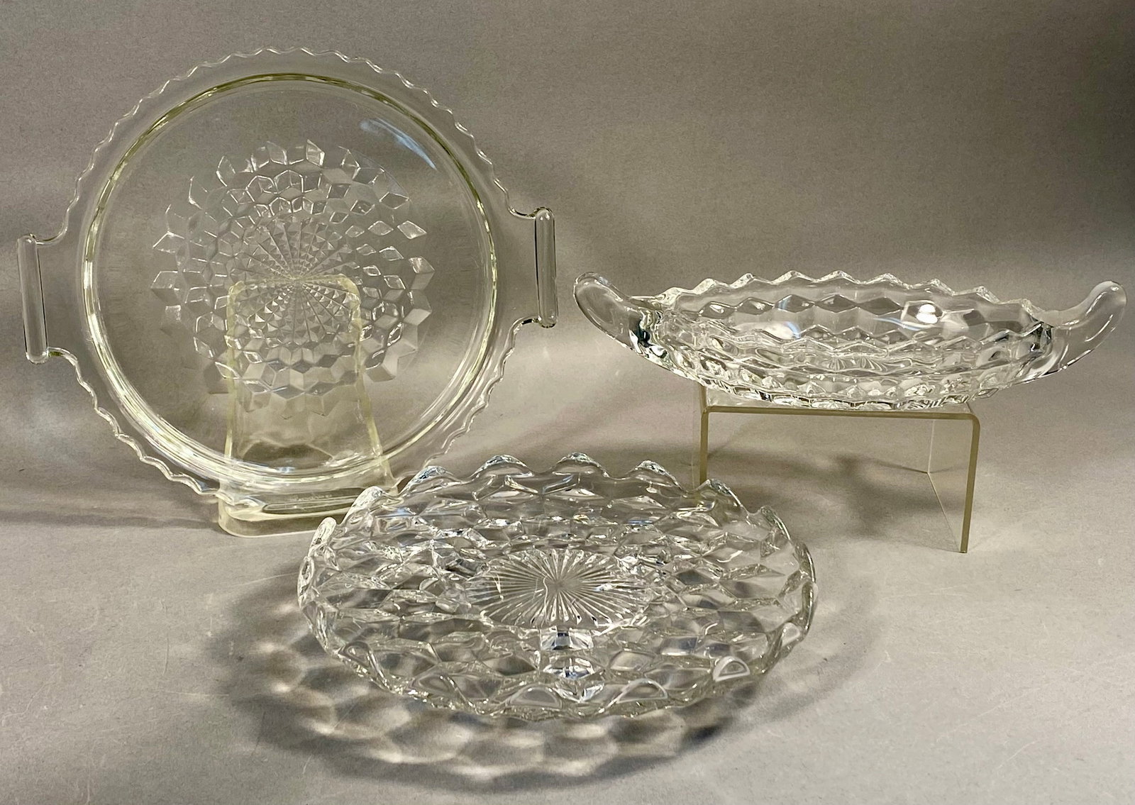A Trio of Fostoria American Pattern Pressed Glass Tableware: This collection consists of three pieces of clear pressed glass in the American pattern, Line 2056, produced by the Fostoria Glass Company. The set includes a circular handled serving tray, a shallow