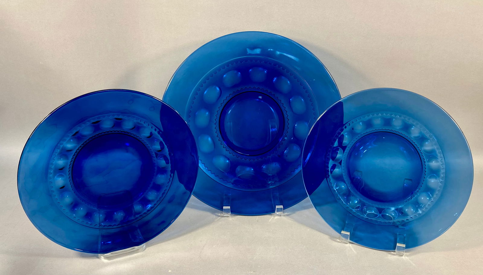 A Trio of Cobalt Blue Glass Plates in the Tiara Crown Imperial Style: This collection includes three cobalt blue pressed glass plates, specifically one large platter and two dinner plates. The pieces feature a decorative pattern consisting of a ring of oval thumbprint i