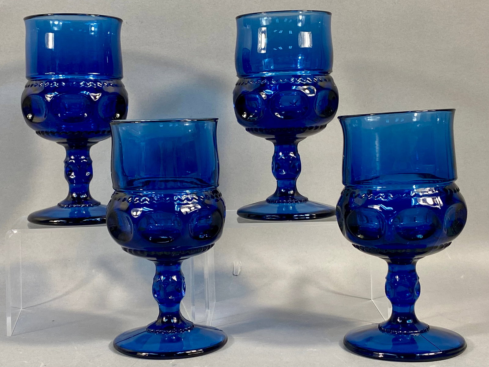 A Set of Four Cobalt Blue Pressed Glass Goblets in the King's Crown Pattern: This set of four pressed glass goblets is rendered in a deep cobalt blue color. The pieces feature the King's Crown pattern, also commonly referred to as the Thumbprint pattern. The design is characte