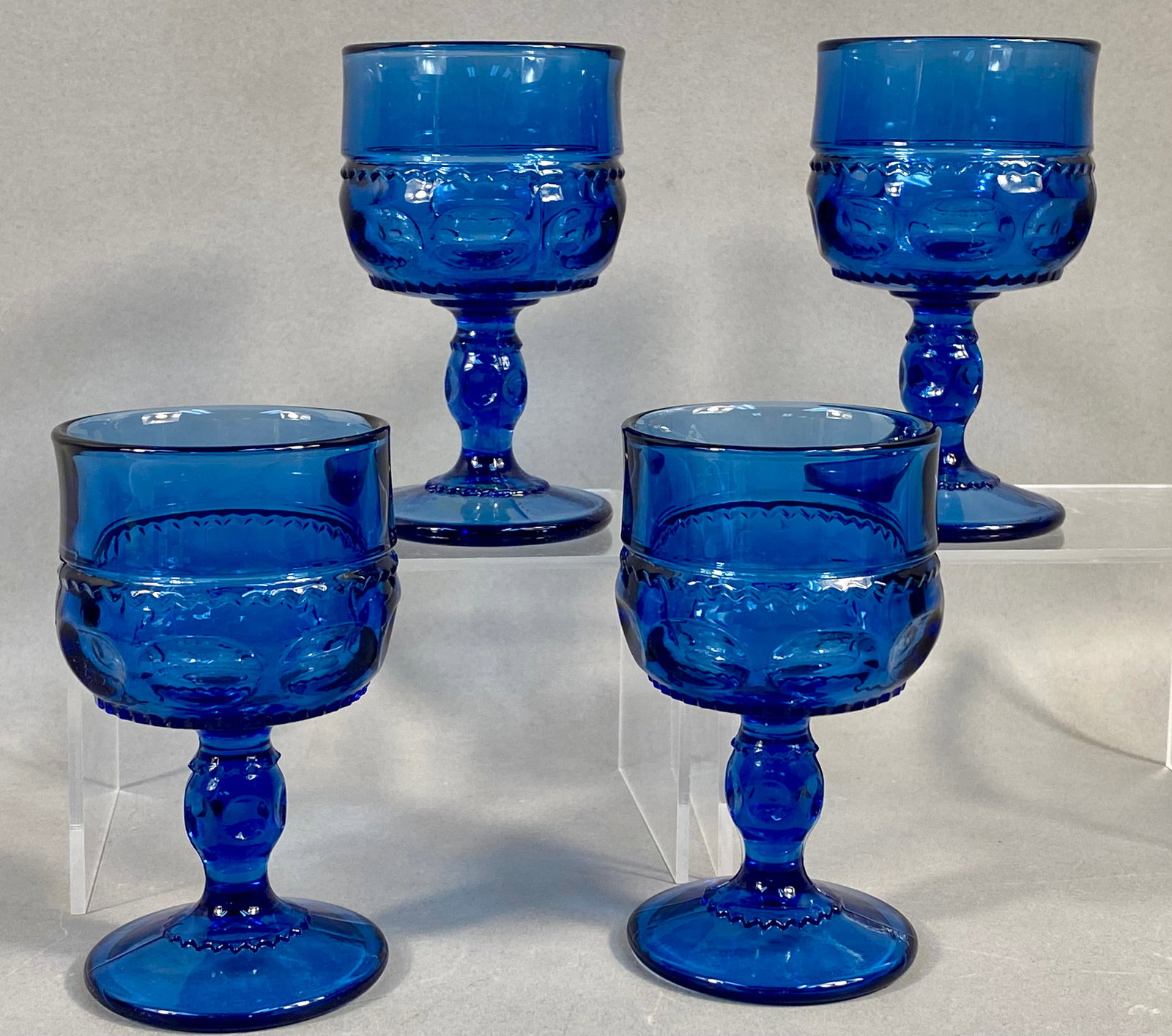 A Set of Four Tiara Exclusive King's Crown Cobalt Blue Glass Goblets: A set of four cobalt blue pressed glass goblets in the King's Crown pattern, also known as Crown Imperial or Tiffin-style Thumbprint. The design features a smooth cylindrical upper rim above a decorat