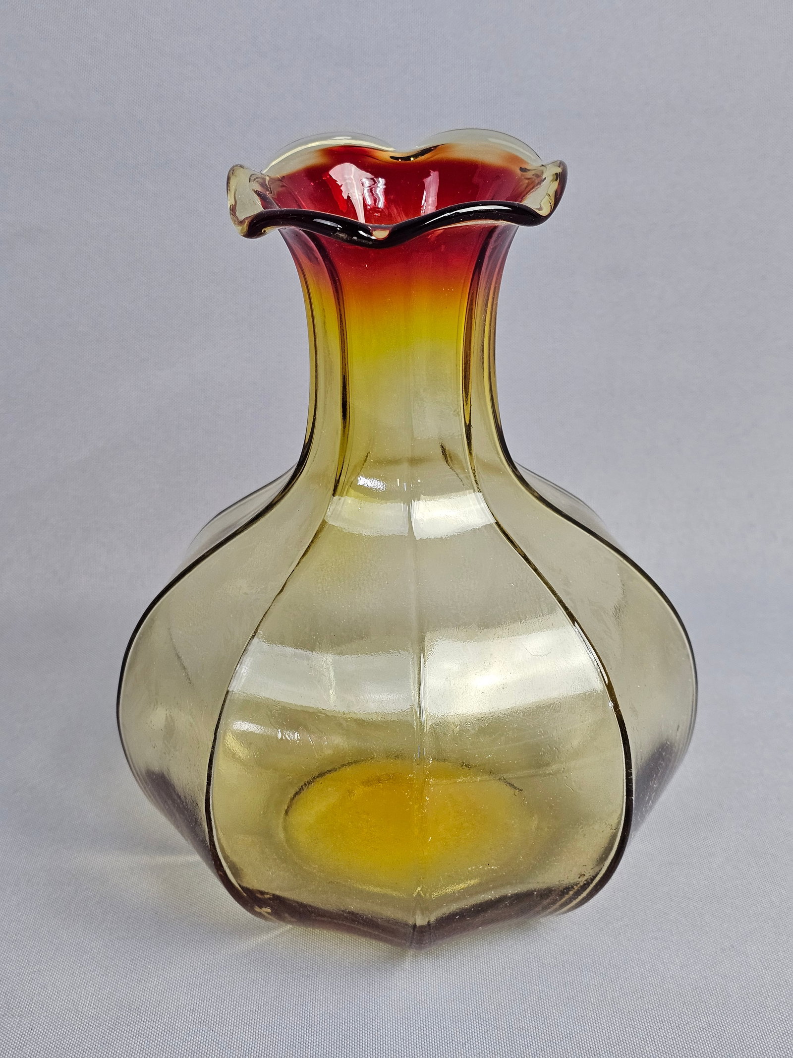 Mid-Century Modern Amberina Art Glass Ribbed Vase Attributed to Blenko: This mid-century modern art glass vase is crafted in the Amberina style, featuring a color gradient that transitions from a yellow-amber base to a ruby red neck and rim. The vessel is designed with a