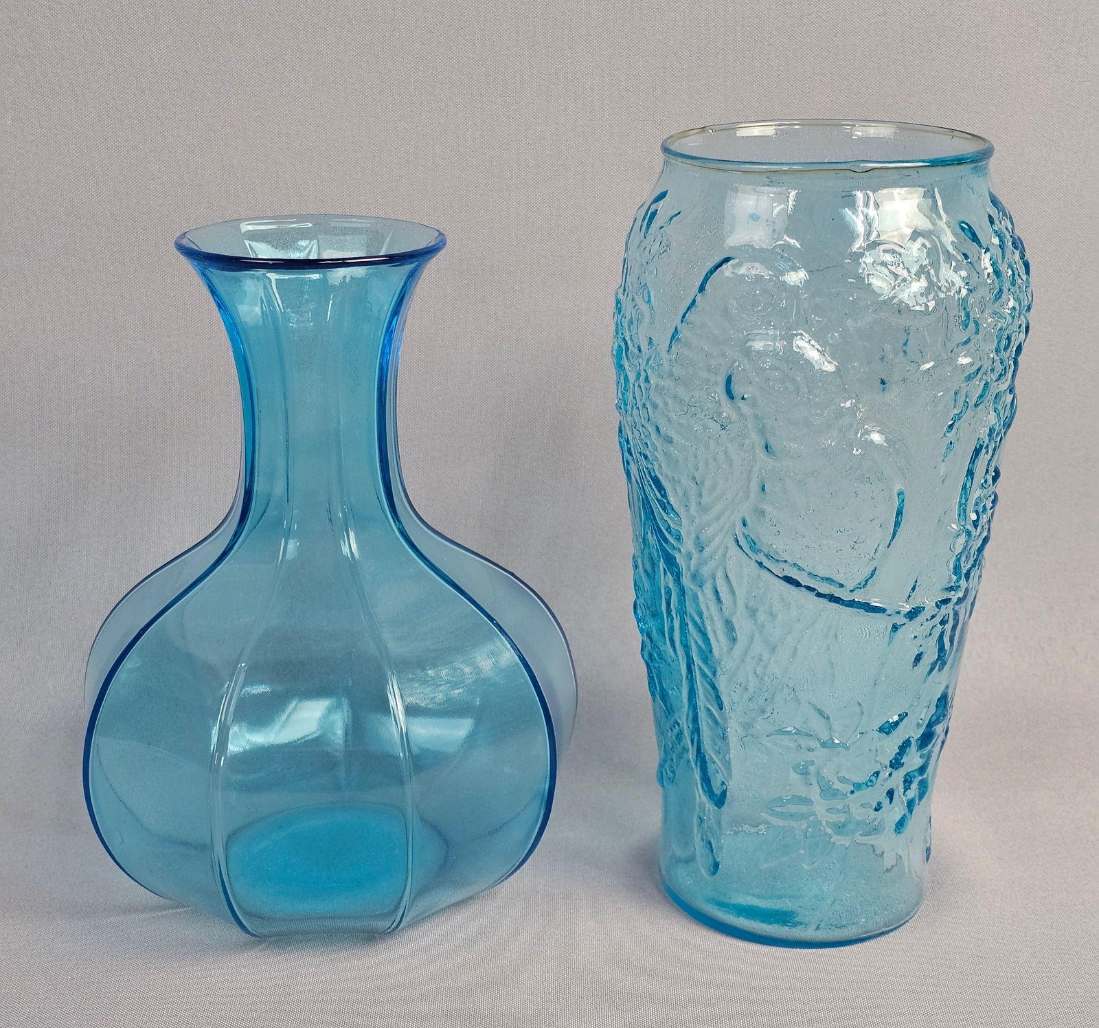 A Pair of Tiara Exclusive Aqua Blue Glass Vases: This lot comprises two decorative glass vases in varying shades of aqua blue. Vases measure 8 and 9 inches tall. Both vases are in good condition with no visible chips, cracks, or repairs. Minor shelf