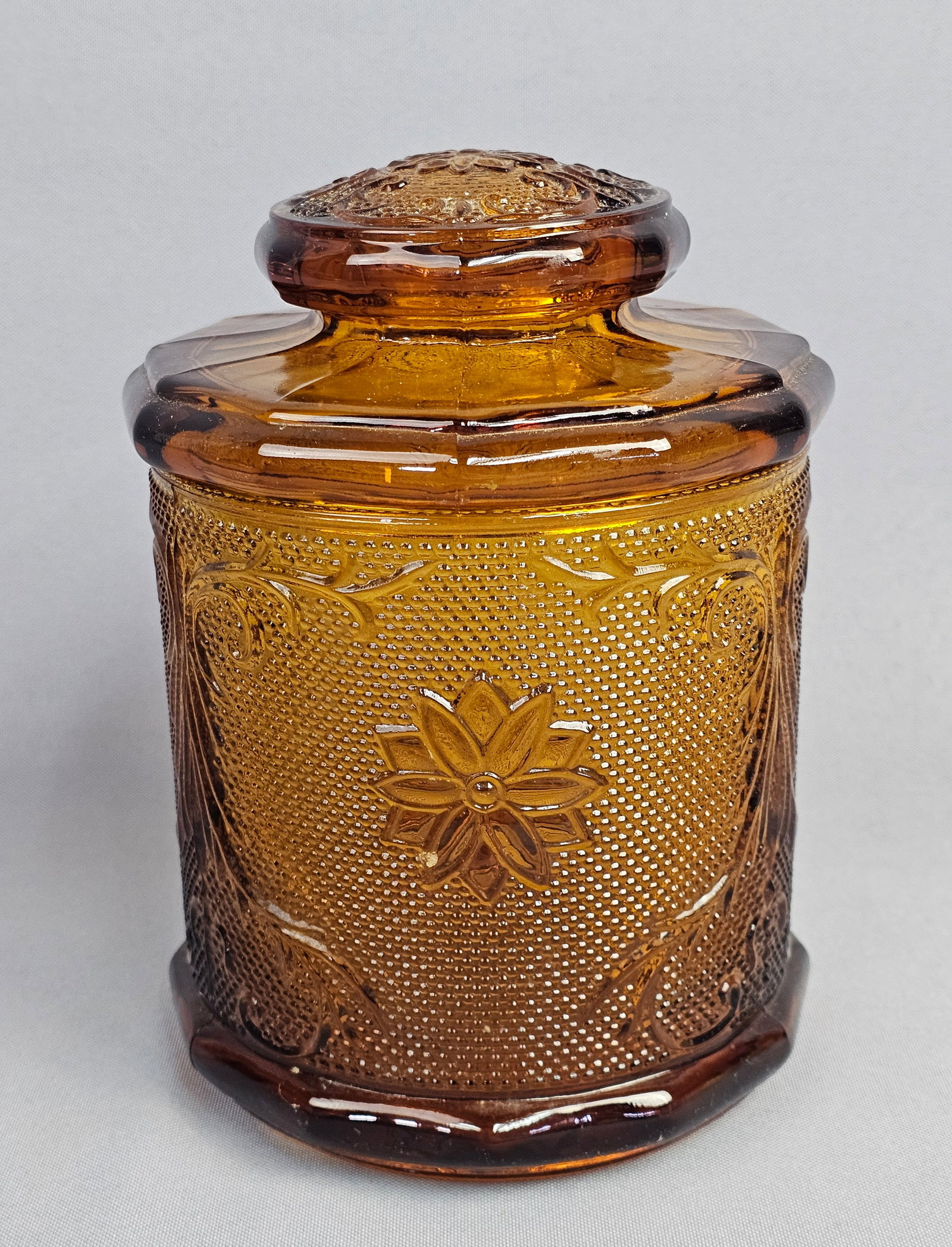 Tiara Indiana Glass Sandwich Pattern Amber Glass Canister: This is a vintage pressed glass covered canister in an amber color. The piece features the Sandwich pattern, characterized by embossed floral starbursts and scrolling vine motifs set against a stipple
