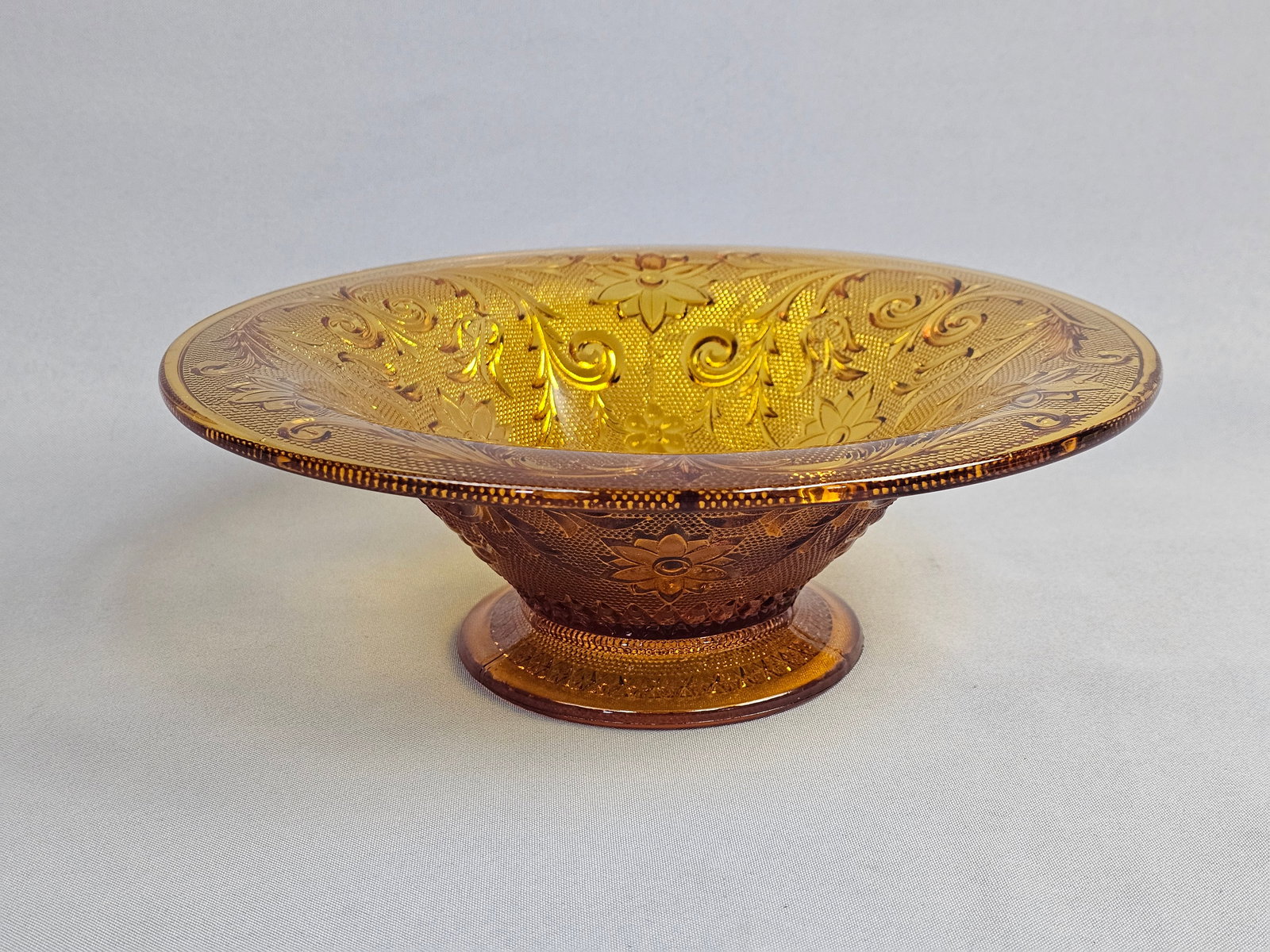 Indiana Glass Tiara Sandwich Pattern Amber Glass Footed Bowl: This footed bowl is produced in the Tiara Sandwich pattern by the Indiana Glass Company, typically dating to the 1970s or 1980s. The piece is made of pressed amber glass with a translucent golden brow