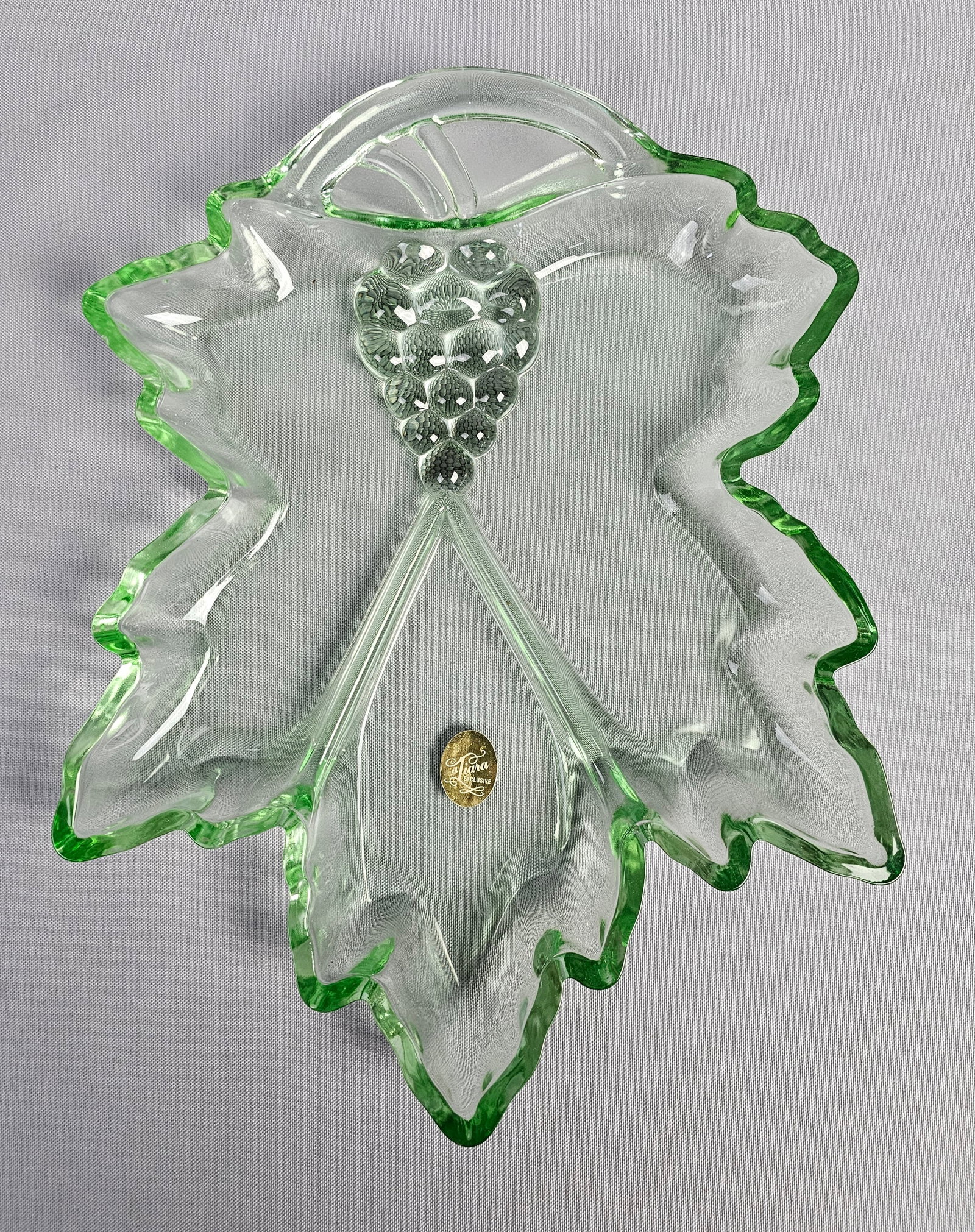 Tiara Exclusives Light Green Pressed Glass Grape Leaf Serving Tray: This is a pressed glass serving tray or decorative dish in the form of a leaf, featuring a transparent light green color. The design includes embossed veining throughout the body and a textured cluste