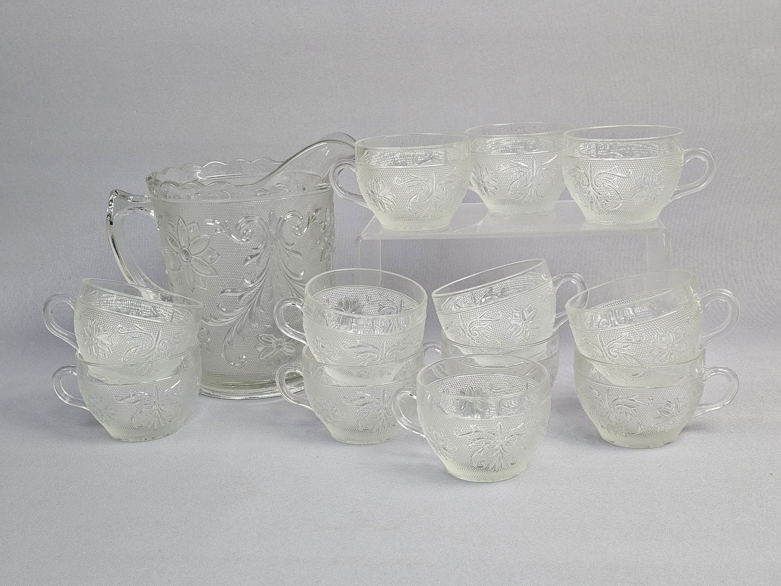 A 13-Piece Set of Clear Pressed Glass Beverage Ware in the Sandwich Pattern: This thirteen-piece clear pressed glass beverage set features the Sandwich pattern, a design widely produced by American manufacturers such as the Indiana Glass Company and Tiara Exclusives. The set c