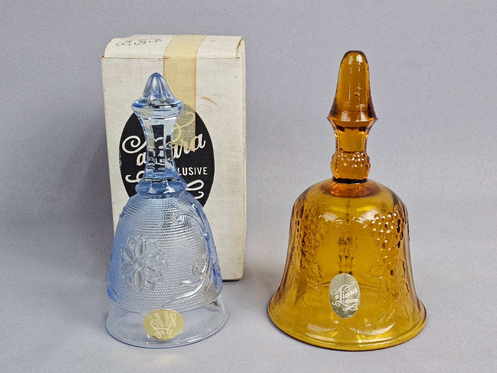 A Pair of Tiara Exclusives Pressed Glass Bells in Harvest and Sandwich Patterns: This pair of collectible pressed glass bells was marketed by Tiara Exclusives and produced by the Indiana Glass Company during the late 20th century. The first bell is crafted from transparent amber g