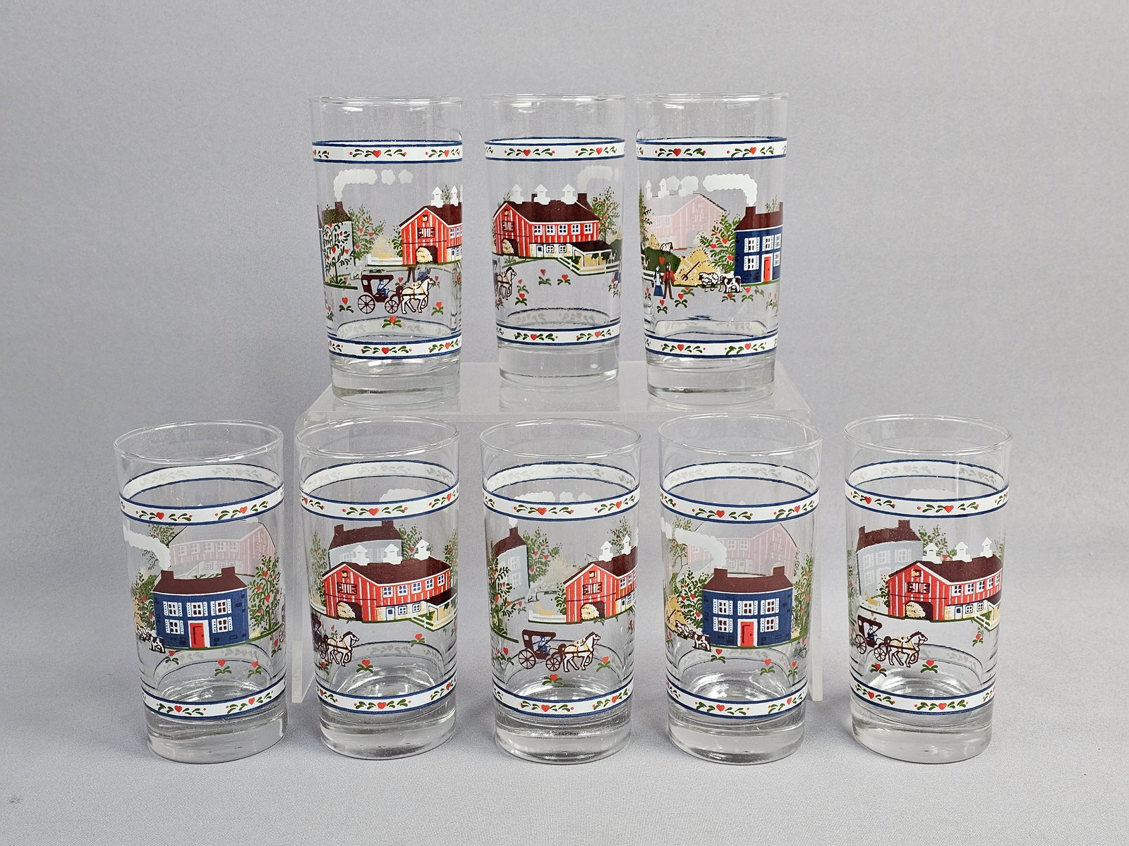 A Set of Eight Tiara Exclusives Heartland Village Pattern Glass Tumblers: This set of eight clear glass highball tumblers features the Heartland Village pattern from the Tiara Exclusives line. Each glass is decorated with a wraparound Americana folk art scene executed in ap