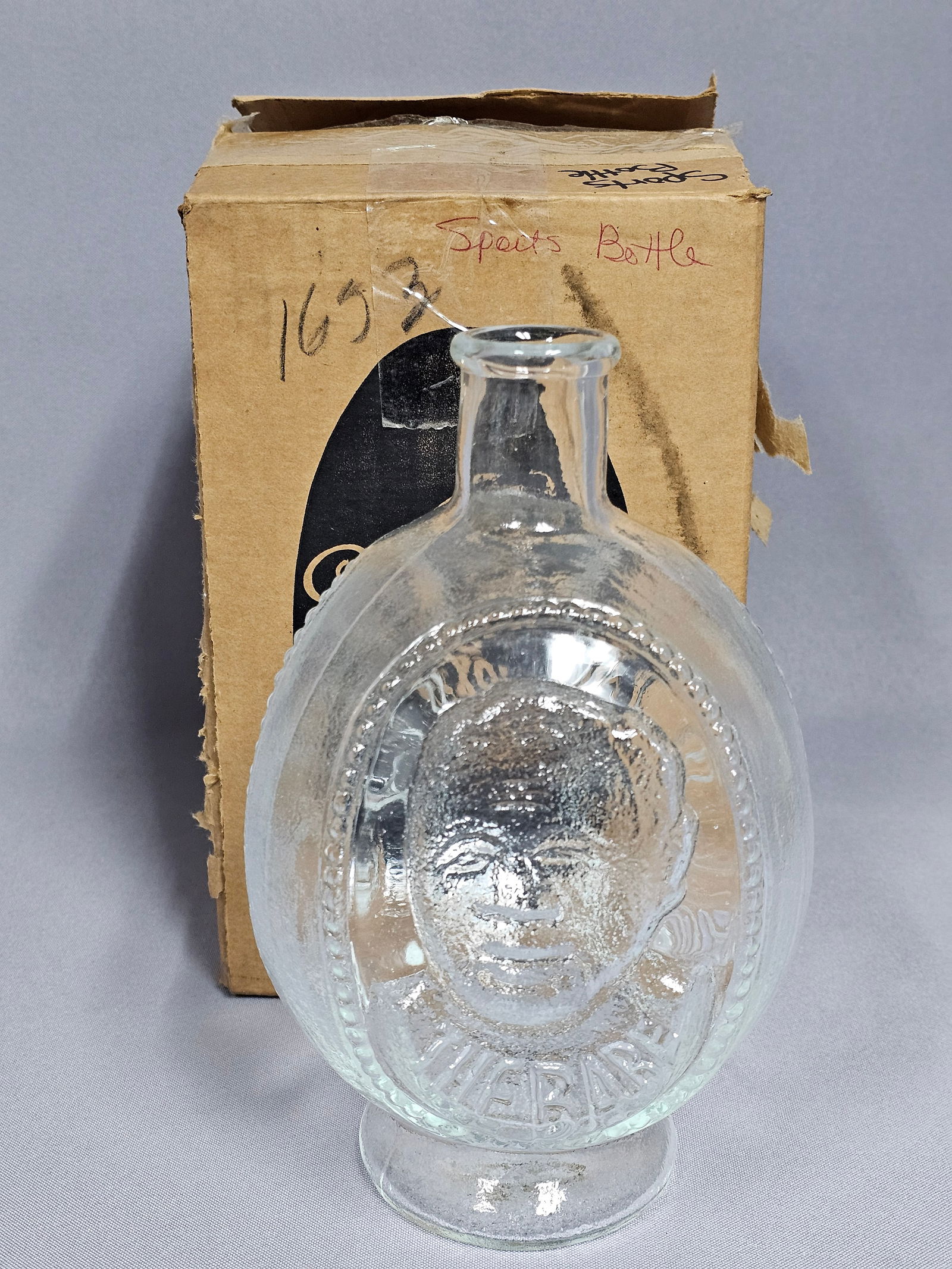 Tiara Exclusives Jim Thorpe Commemorative Pressed Glass Sports Bottle with Original Box: A clear pressed glass commemorative bottle manufactured by Tiara Exclusives, featuring the American athlete Jim Thorpe. The vessel is designed in a flattened oval form with a short neck and flared rim