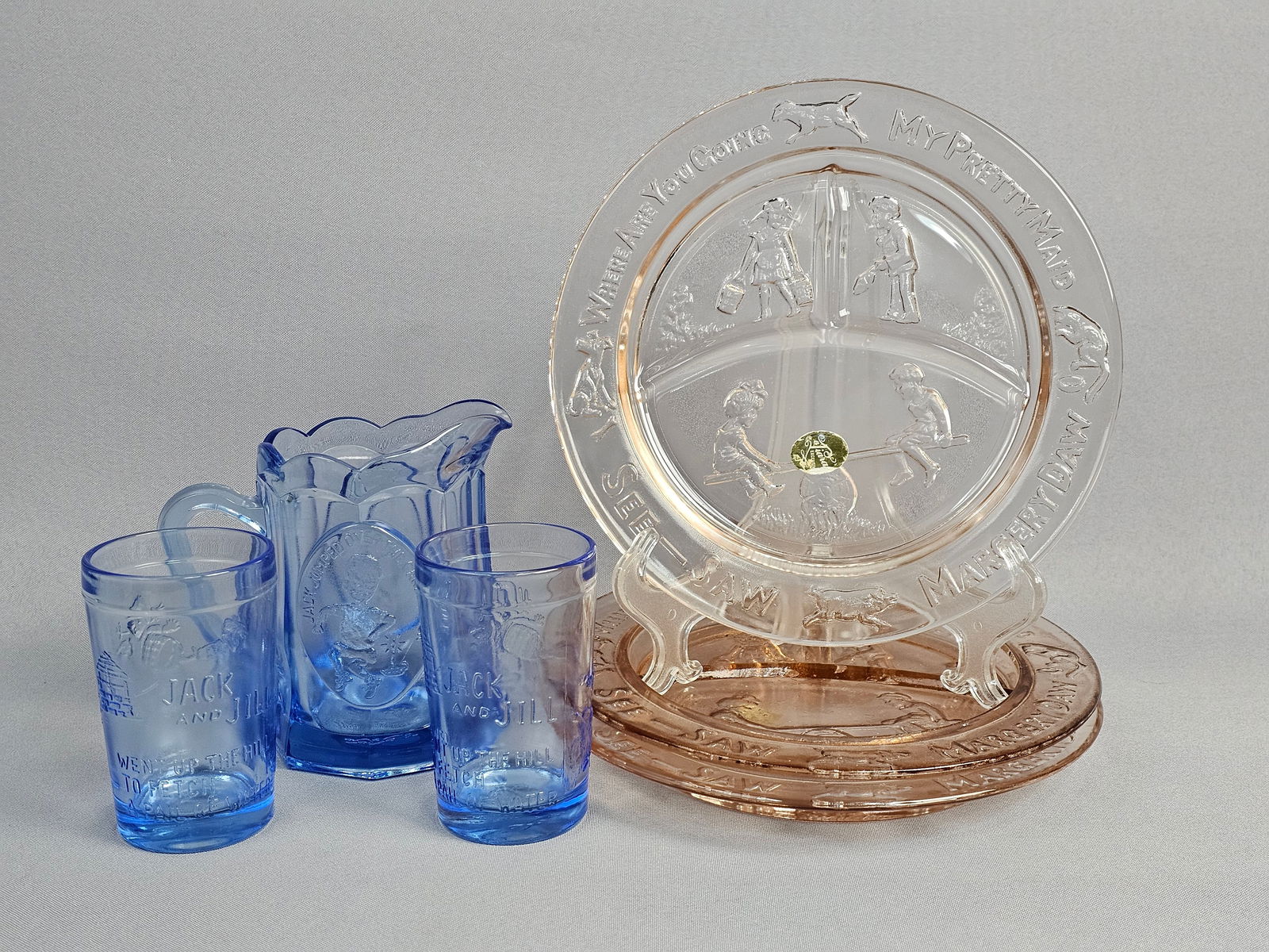 A Collection of Tiara Exclusives Nursery Rhyme Pattern Childrens Glassware in Pink and Blue: This collection of pressed glass children's dinnerware is produced in the Nursery Rhyme pattern by Tiara Exclusives. The assortment includes translucent pink and blue pieces featuring embossed charact