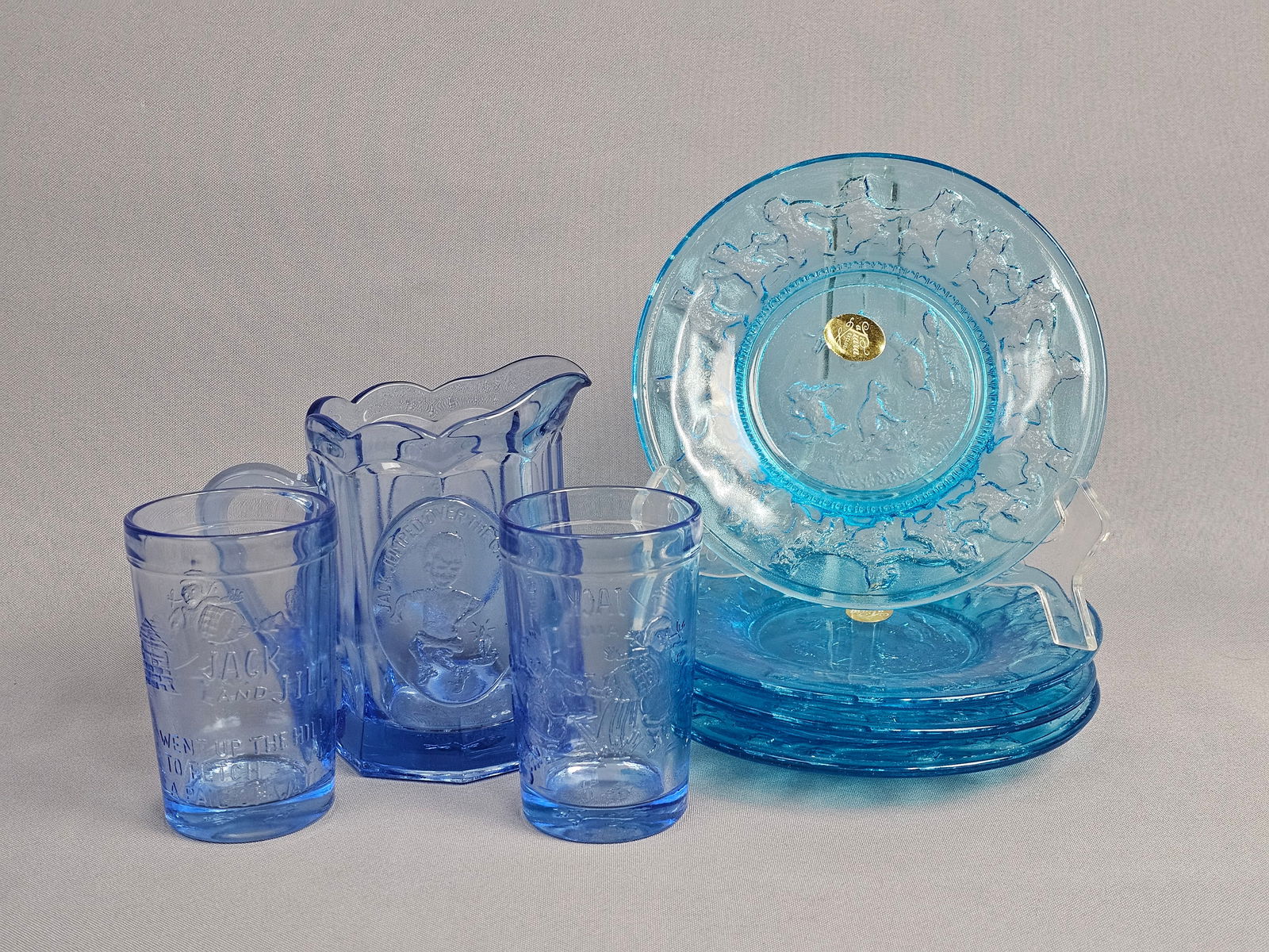Tiara Exclusives Blue Pressed Glass Nursery Rhyme Child's Tableware Set: A seven-piece child's nursery rhyme tableware set produced by Tiara Exclusives in transparent light blue pressed glass. The set comprises a small pitcher, two tumblers, and four small plates. The pitc