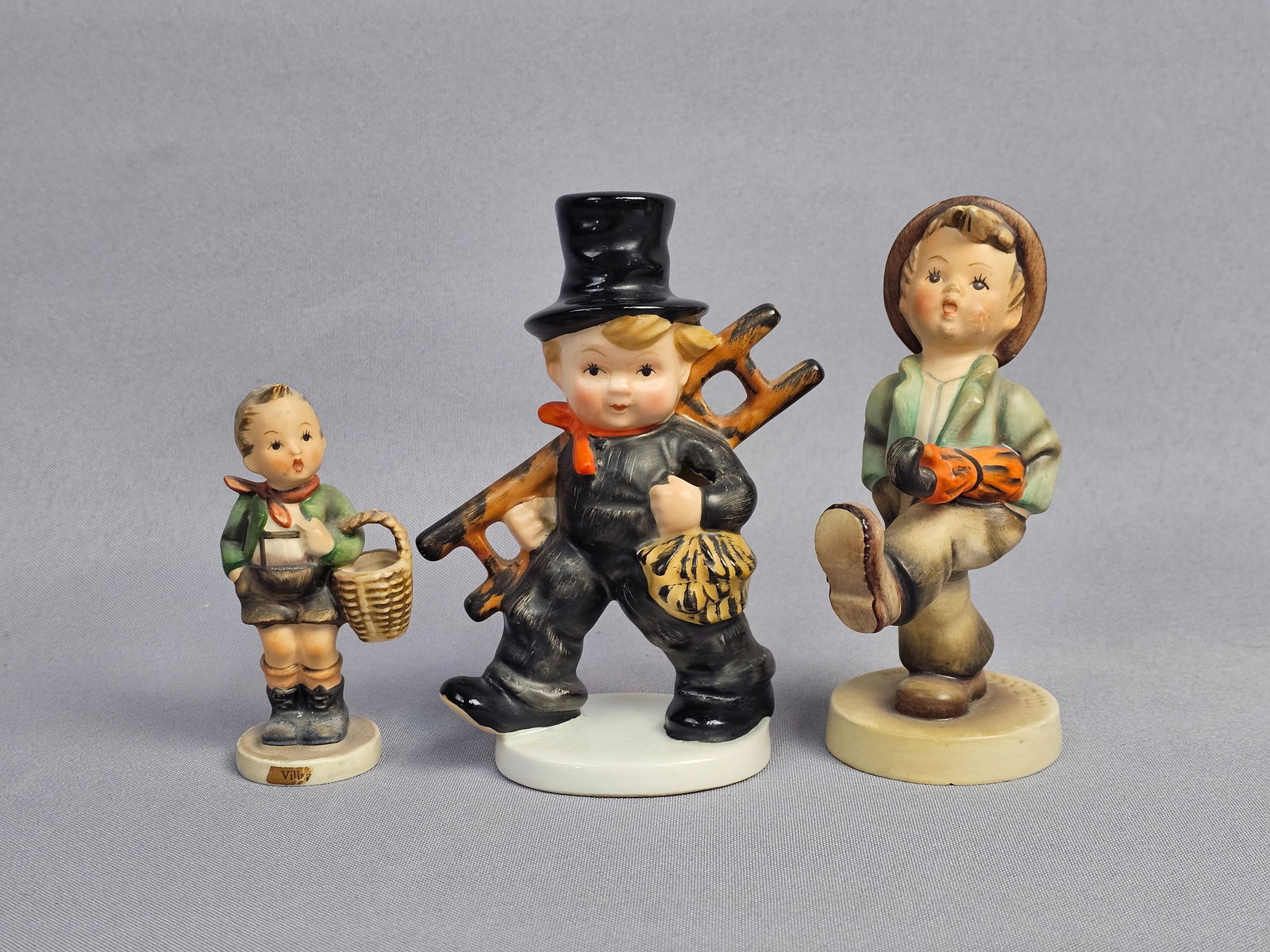 Three Goebel Hummel Hand Painted Ceramic Figurines including Chimney Sweep: This collection consists of three hand-painted ceramic figurines produced in Germany by Goebel in the M.I. Hummel style. The first figurine represents a chimney sweep in a black suit and top hat with