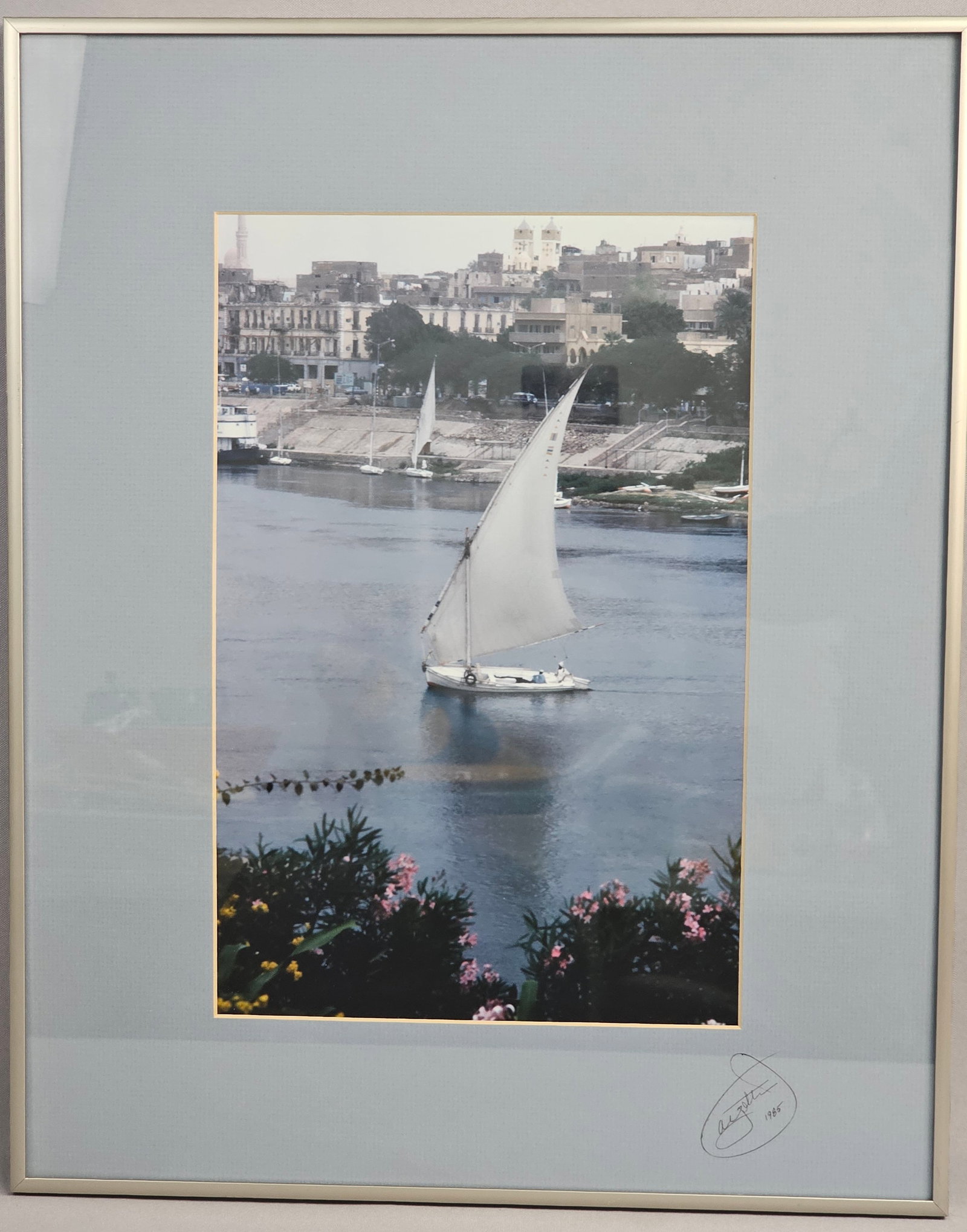 James E. Ayotte, Felucca, Aswan, Egypt, Chromogenic Print, 1985: This chromogenic color print by James E. Ayotte depicts a traditional felucca sailing on the Nile River with the city of Aswan, Egypt, in the background, framed by blooming pink flowers in the
