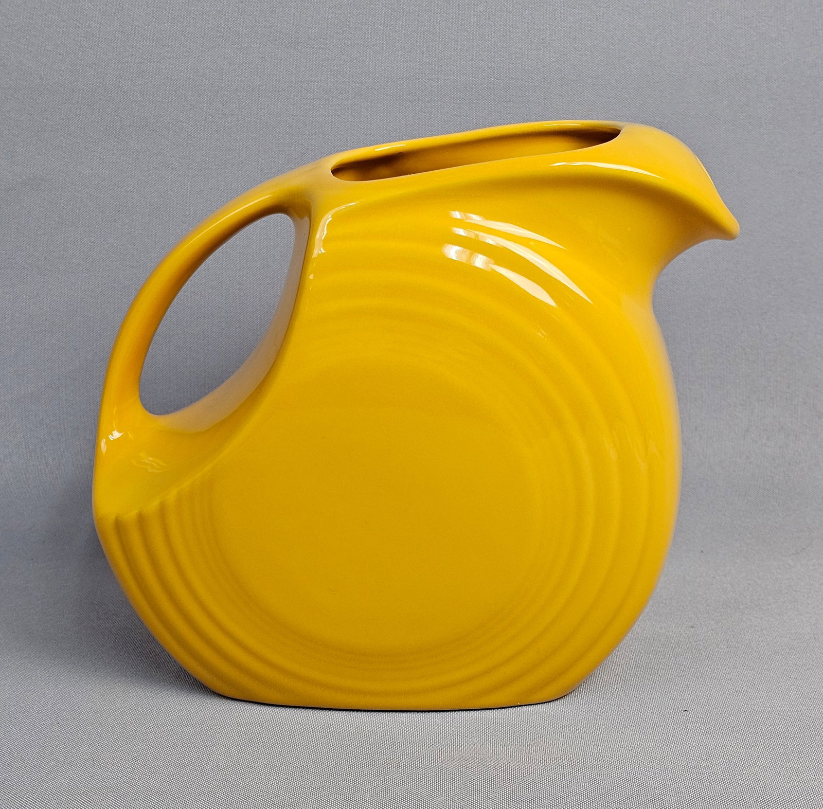 Homer Laughlin Fiesta Yellow Ceramic Disk Water Pitcher: This ceramic pitcher is a product of the Homer Laughlin China Company from the Fiesta dinnerware line. Known as a disk water pitcher, the form features a flattened circular body with an integrated han