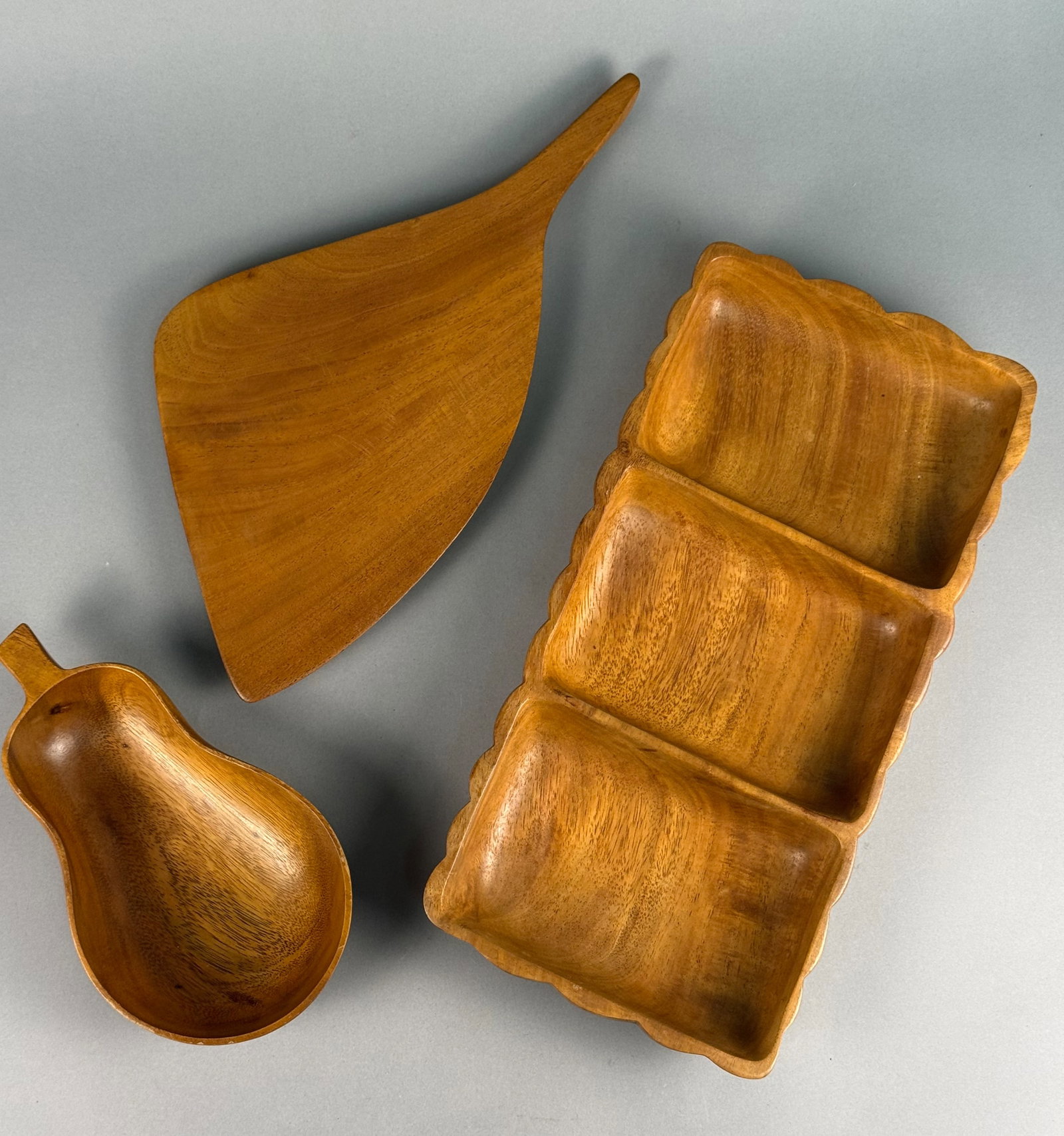 A Trio of Hand-Carved Monkey Pod Wood Serving Vessels: This group of hand-carved monkey pod wood serving vessels features a Mid-Century Modern aesthetic. The collection includes a rectangular tray with three divided compartments and a scalloped rim, a sha