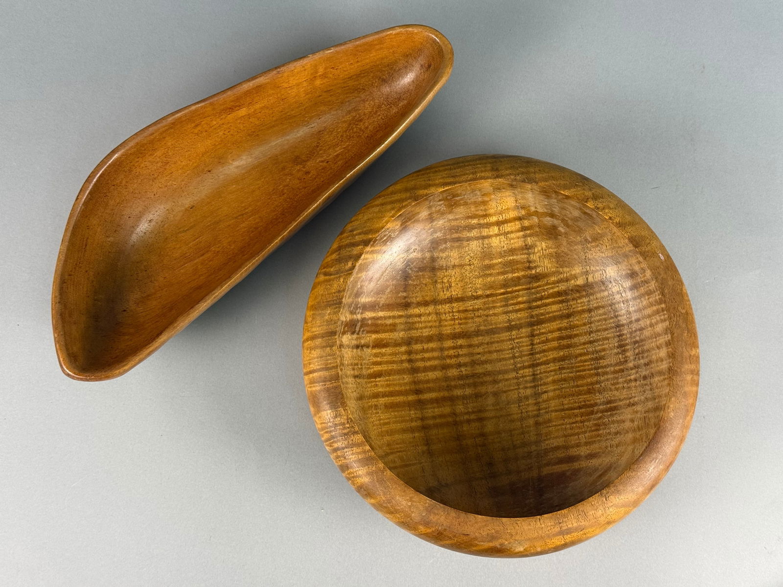 A Pair of Mid-Century Modern Monkey Pod Wood Bowls: A pair of mid-century modern vessels carved from monkey pod wood. The first is a circular hand-turned bowl featuring a tiger grain pattern, a rounded rim, and a raised circular foot. The second is an