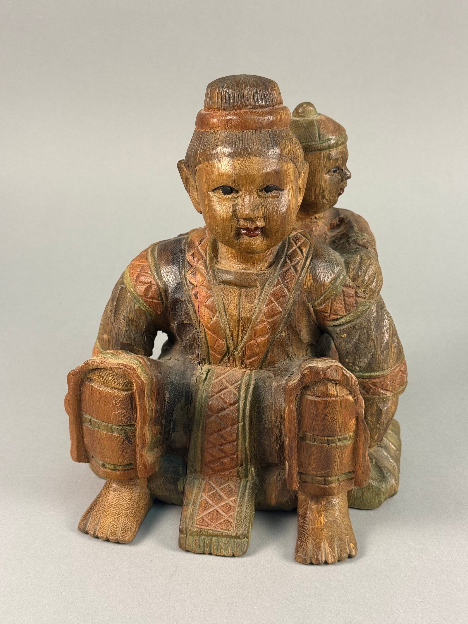 Southeast Asian Carved Wood Polychrome Sculpture of Two Figures: This hand-carved wooden sculpture depicts two figures in a secular folk scene, featuring a larger crouching adult carrying a smaller child on their back. The figures are rendered in a style characteri