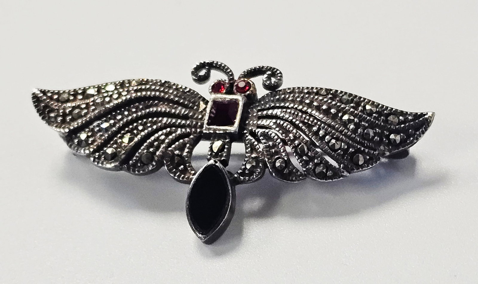 Sterling Marcasite & Garnet Moth Brooch 8.0 Grams: This sterling silver brooch is designed in the form of a luna moth with an oxidized finish. The reverse of the piece is stamped 925. The brooch is secured with a horizontal pin and a rolling safety